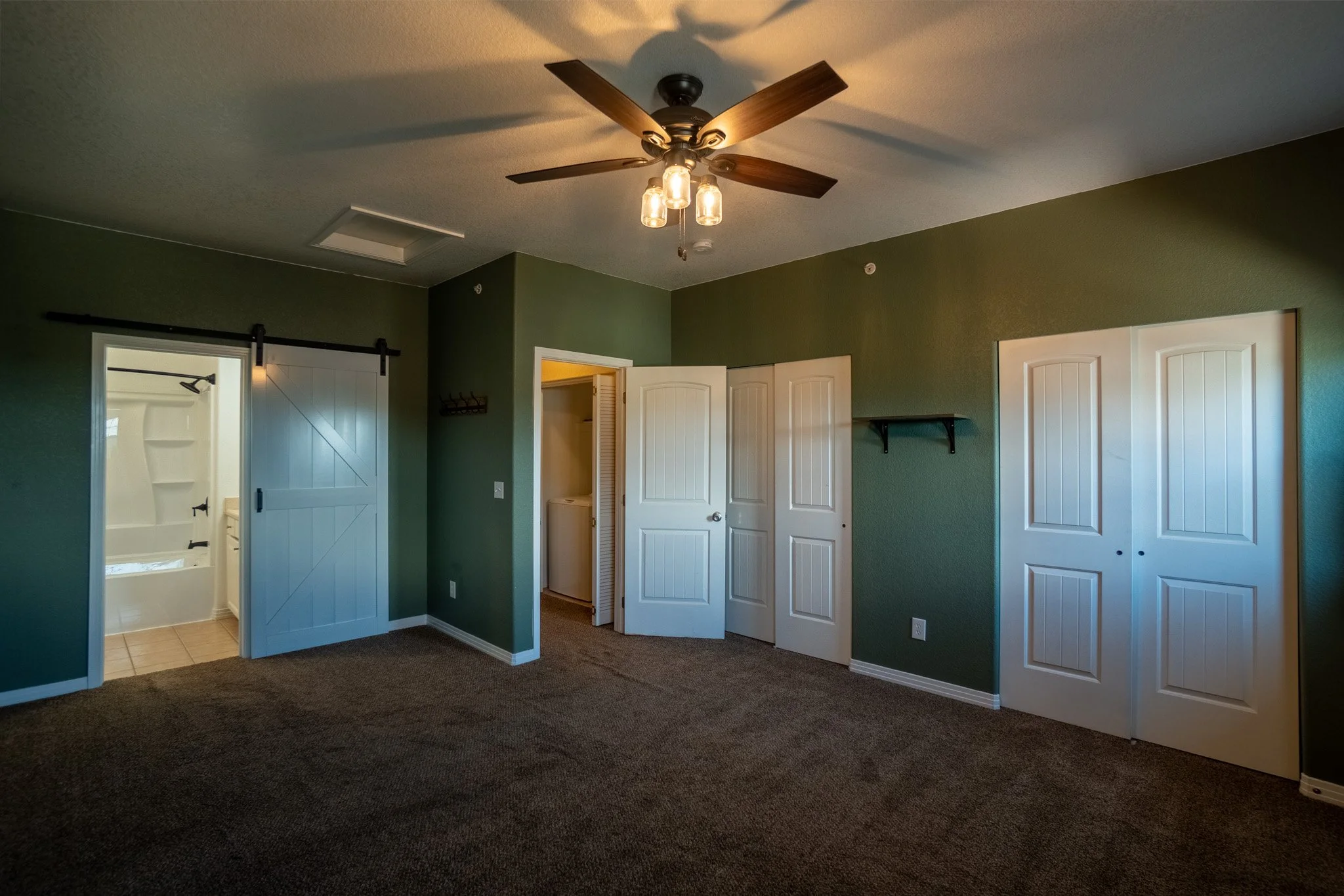 Interior of a room with green walls, white doors, and brown carpet. Ceiling fan with wooden blades and multiple light bulbs. Partial view of a bathroom with a shower.