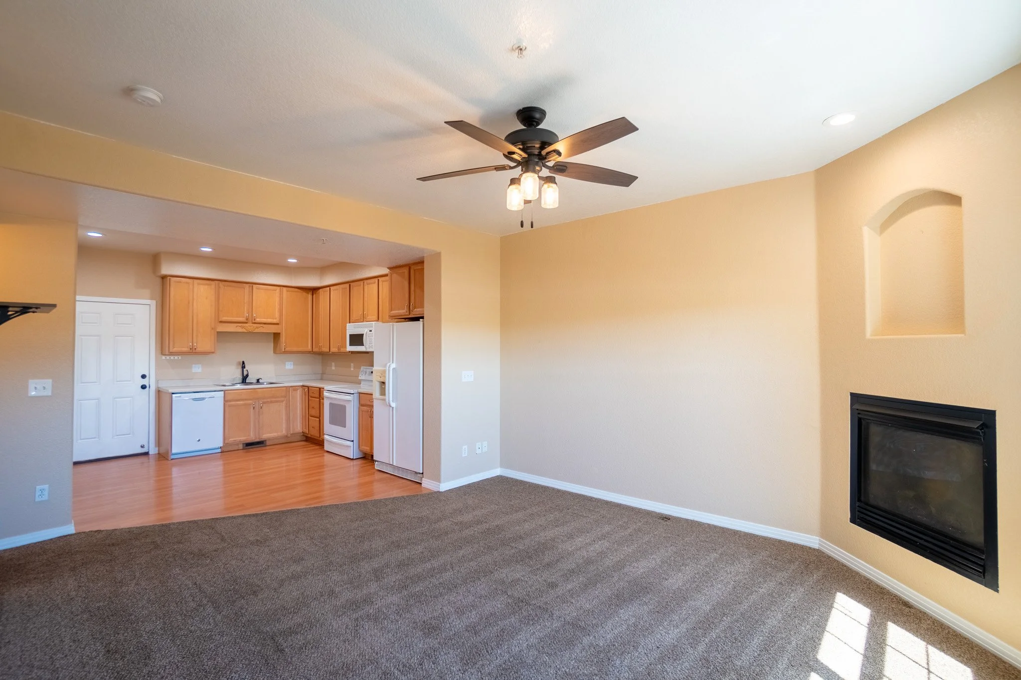 Living area with a fireplace, beige walls, and a ceiling fan with lights. Open view to a kitchen with wooden cabinets and white appliances.
