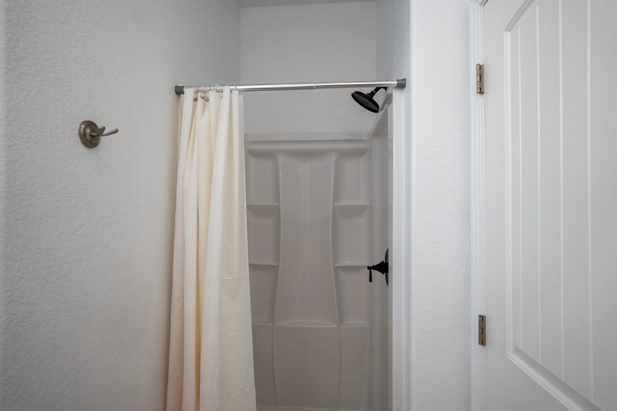 Bathroom shower with beige curtain on a silver rod, white walls, and a built-in shower area with shelves, viewed from the entrance.