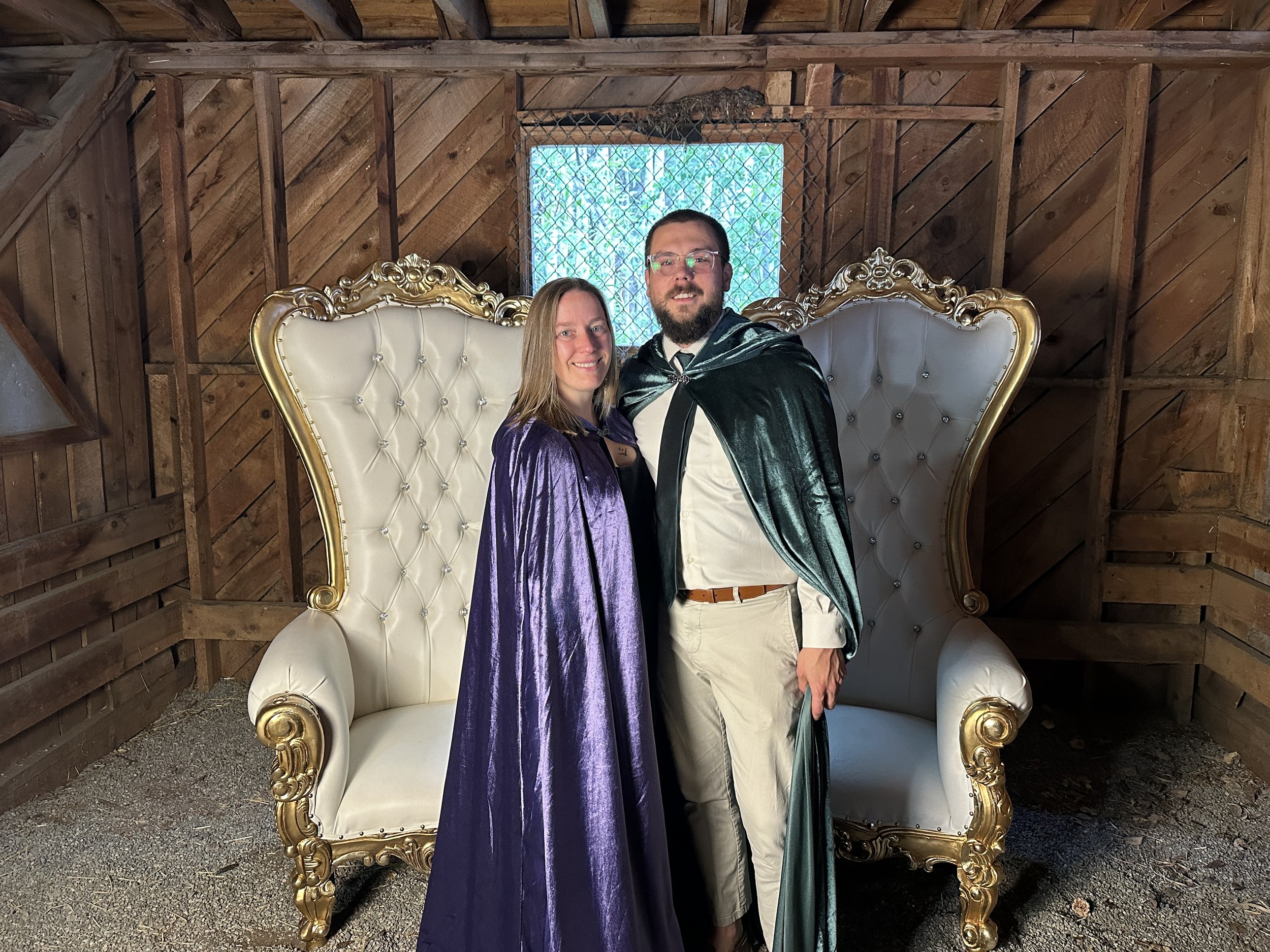 A couple dressed in formal wedding attire sitting on a fancy white and gold throne-like chair inside a wooden structure. There is a small window behind them with a mesh cover, and a small lizard is on a wire above the window.