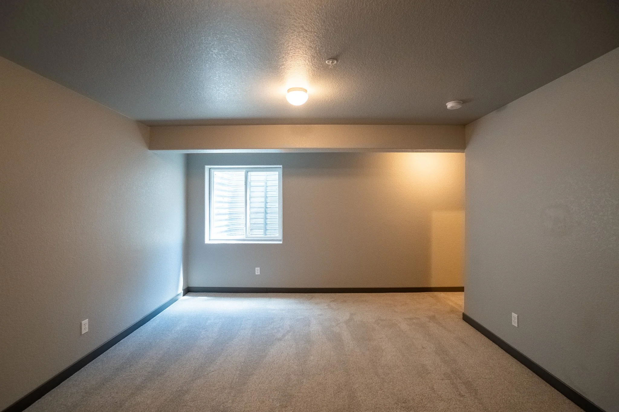 Empty room with beige walls, gray baseboards, window with blinds, ceiling light, and carpeted floor.