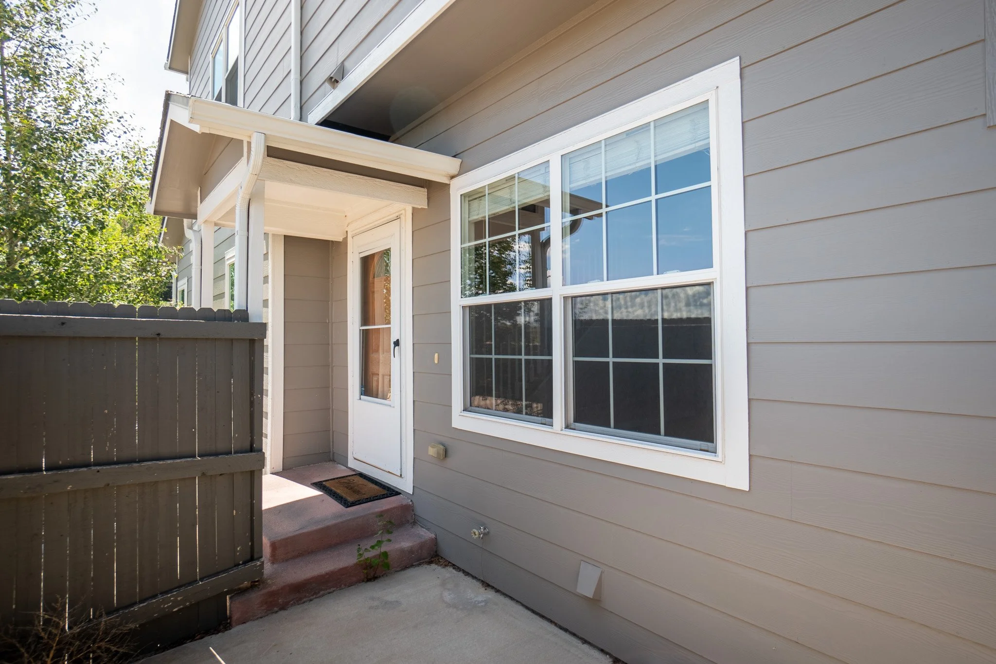 Back entryway of a house with a gray exterior, a white door with a small window, a large window beside it, a small ramp with concrete steps, and a gray fence to the left. There are steps leading up to the door.