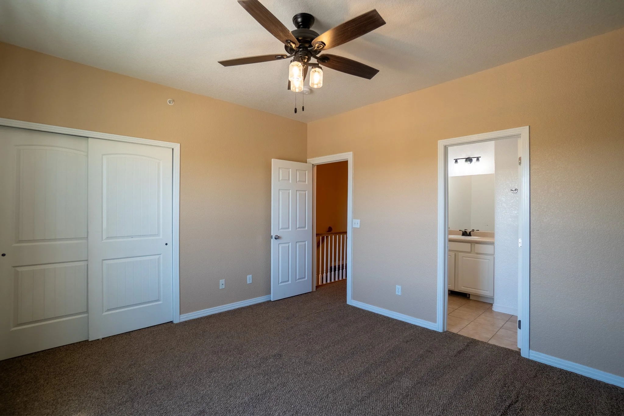 Interior of an empty room with beige walls, ceiling fan, white closet doors, an open door leading to a hallway, and a doorway to a bathroom with a sink.