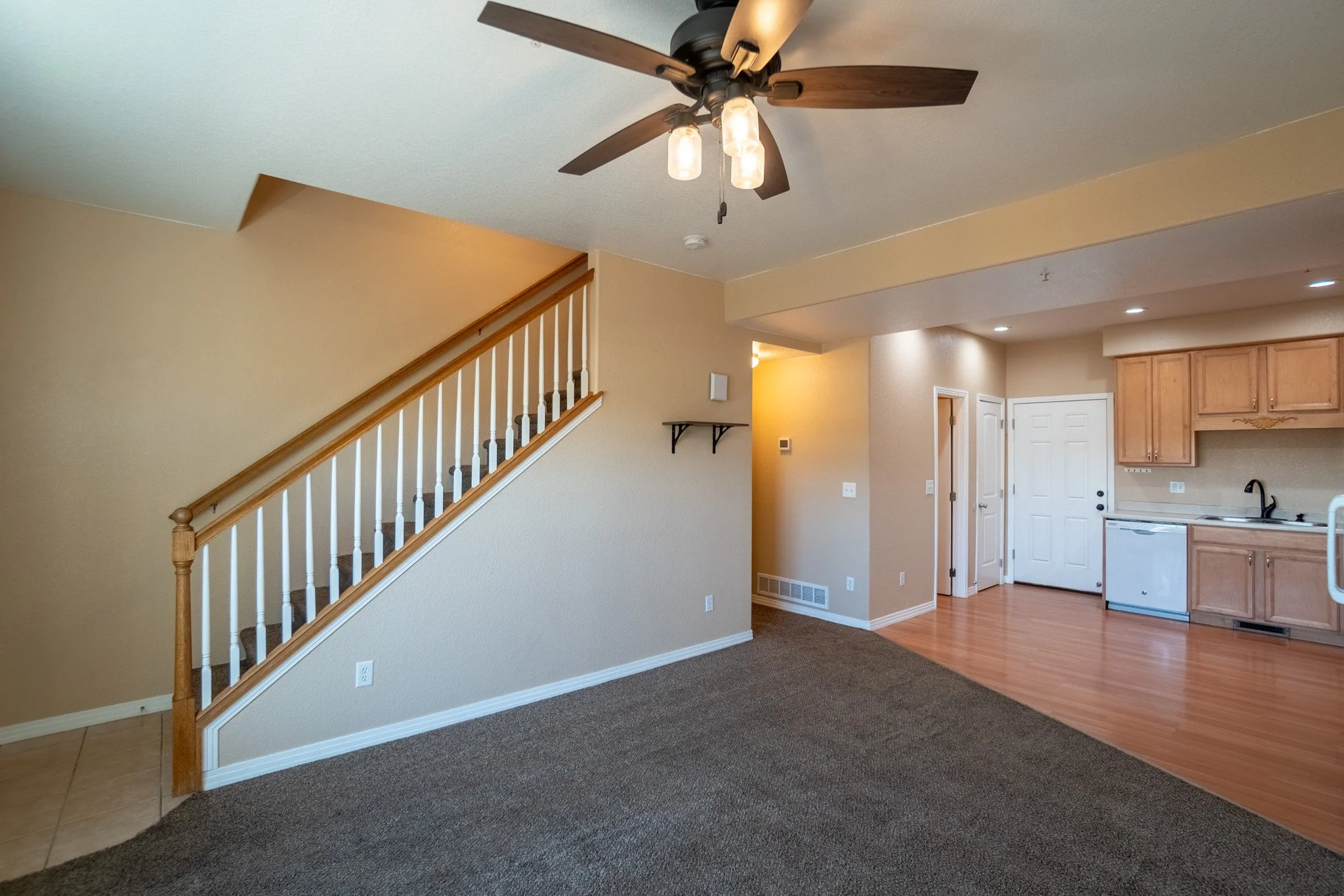 Interior view of a living room and kitchen area in a house, with a staircase, beige walls, wooden cabinets, and a mix of carpeted and hardwood floors.