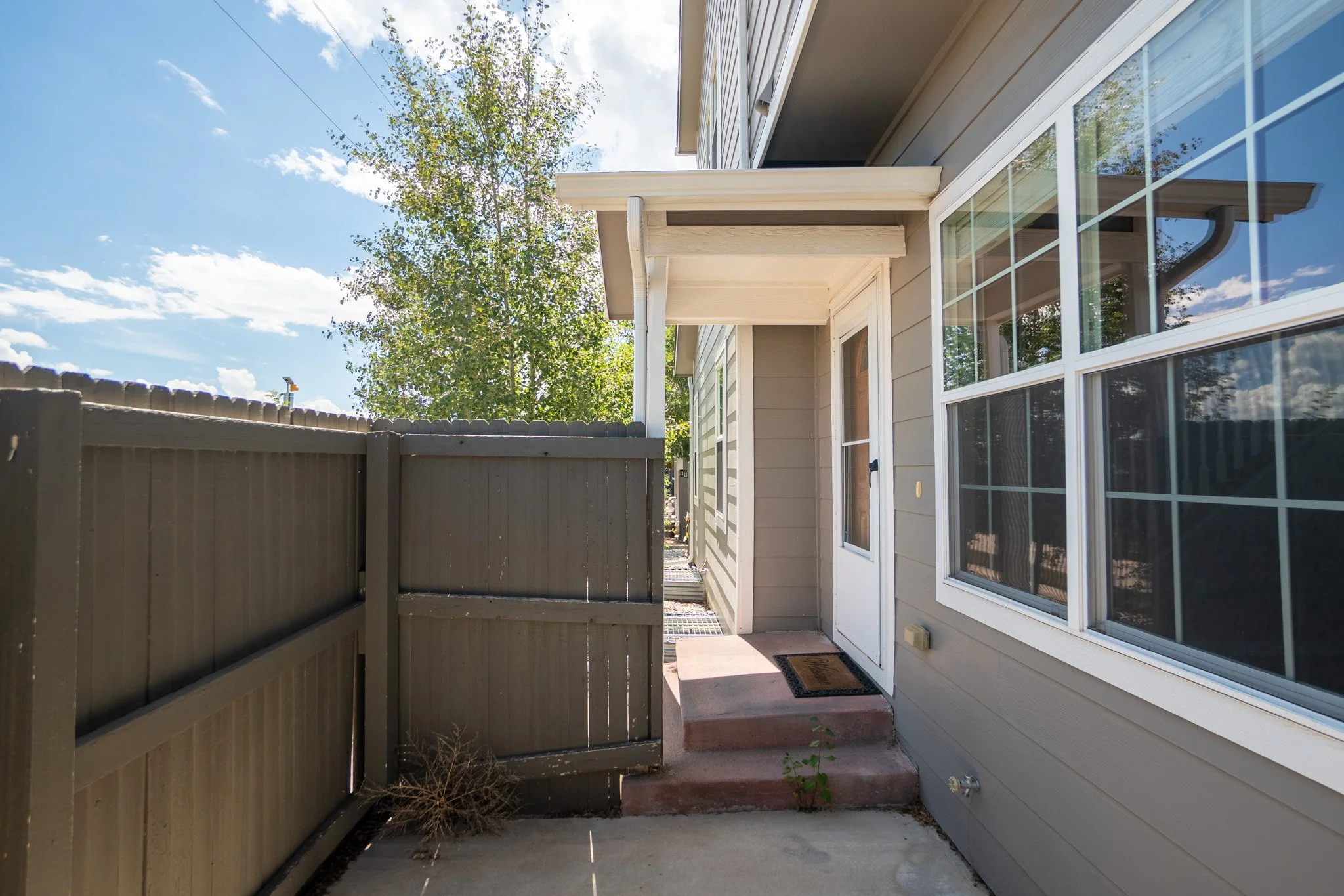Backyard patio with beige fence, concrete ground, and house with large windows and siding, under a partly cloudy sky.