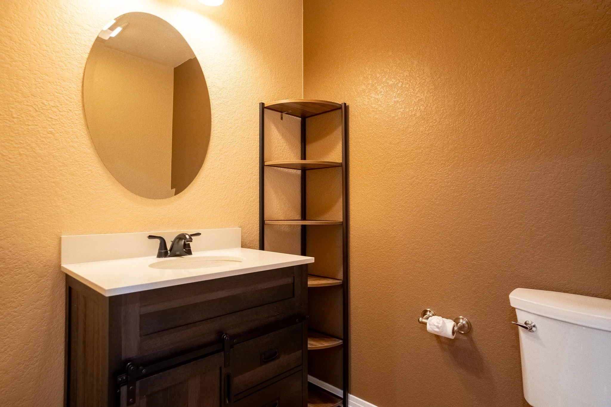 Bathroom with a wall-mounted mirror, a dark wood vanity sink, a metal shelving unit, a toilet with a toilet paper roll, and beige textured walls.