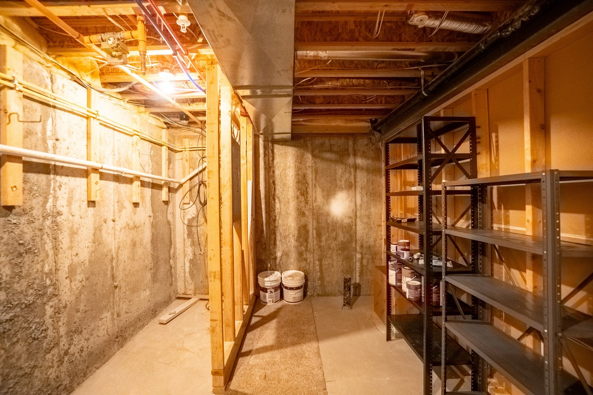 An unfinished basement with exposed wood framing, concrete walls, and a shelving unit holding paint cans, with pipes and electrical wiring overhead.