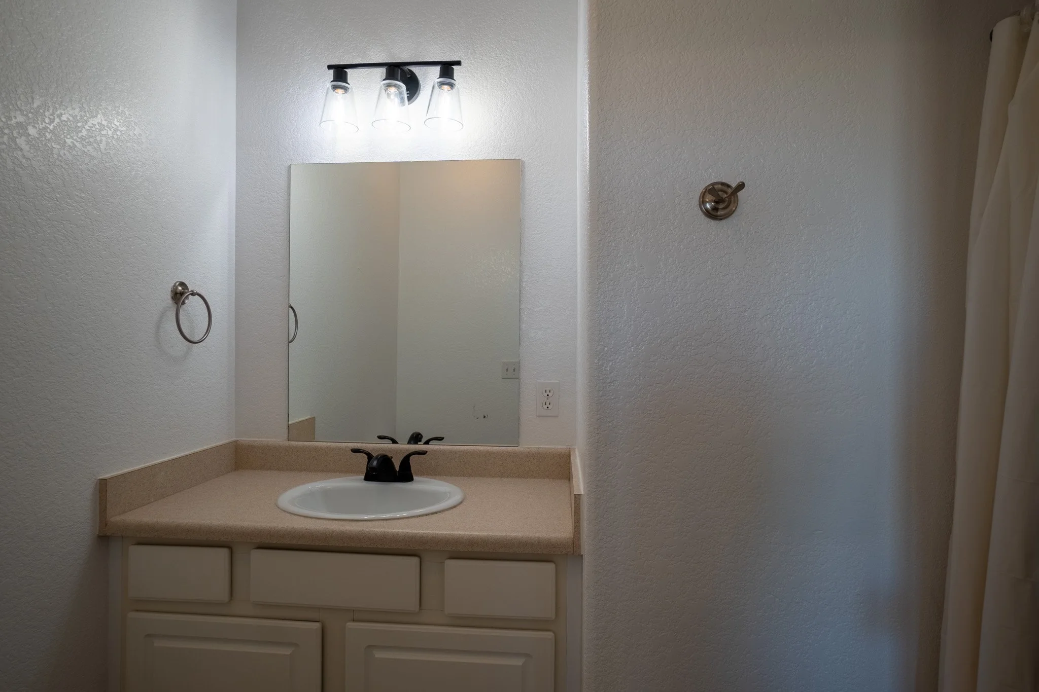 Bathroom vanity with a mirror, black faucet, overhead light fixture with three glass shades, towel ring on the left wall, and beige countertop with a cabinet below.