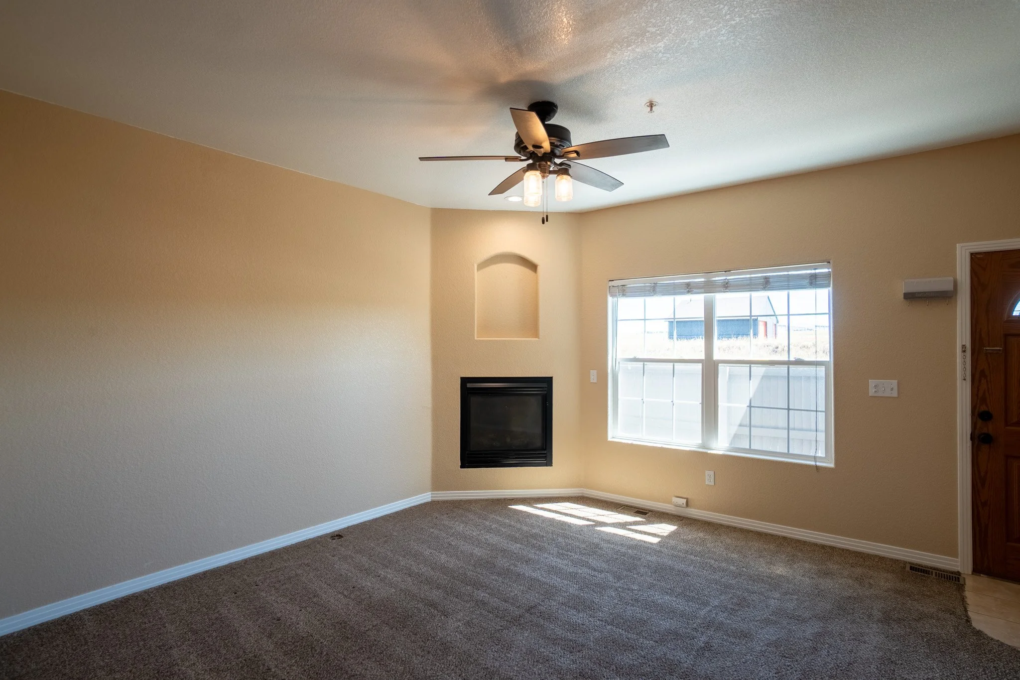 Empty living room with beige walls, brown carpet, a large window with blinds, a fireplace, and a ceiling fan with lights.