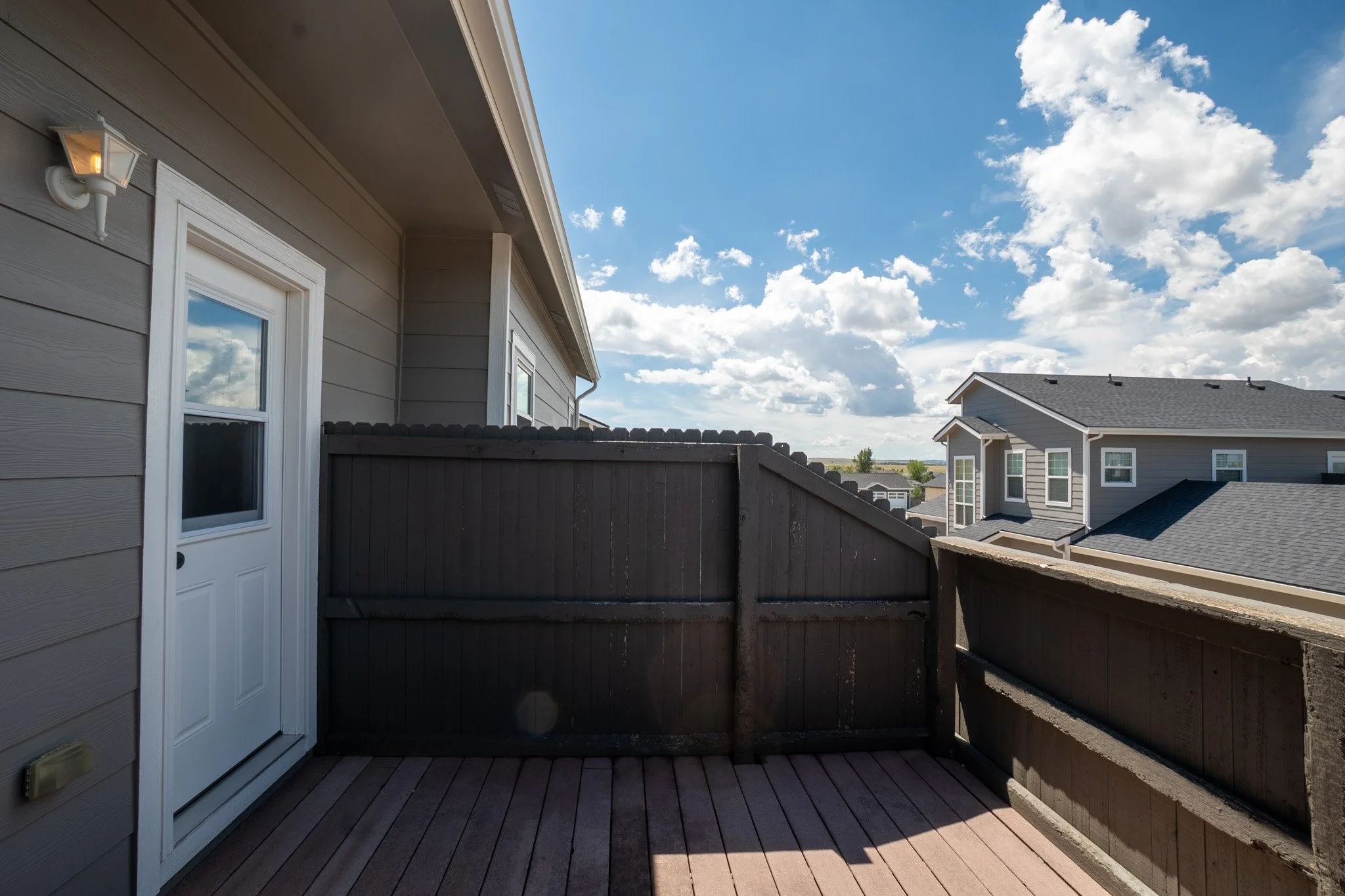 A view from a small fenced backyard balcony with wooden flooring, showing the side of a house with beige siding and a white door and window, under a partly cloudy sky with white fluffy clouds.
