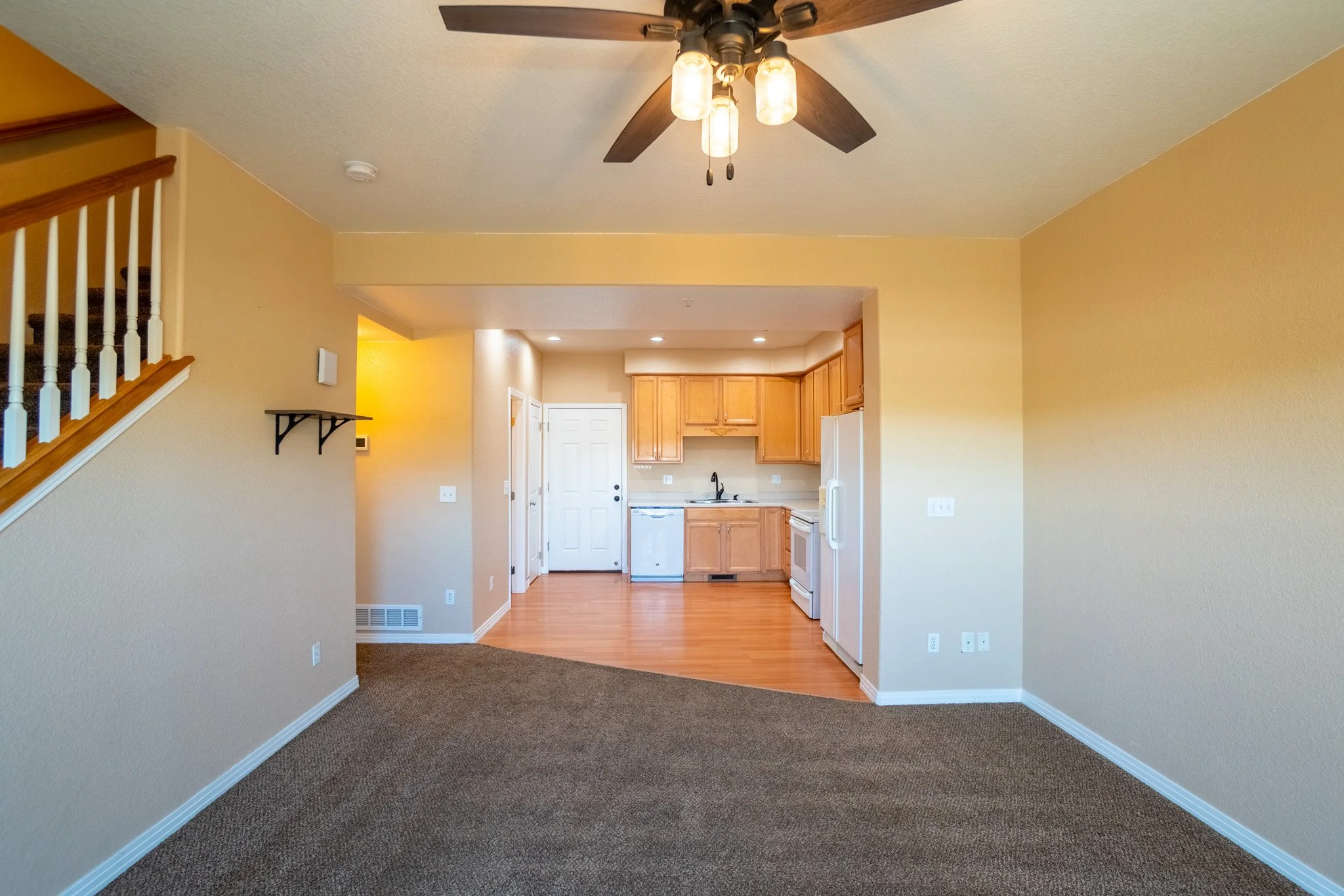 Interior view of a kitchen and living area in a home with yellow walls, wooden cabinets, white appliances, and a ceiling fan with lights.
