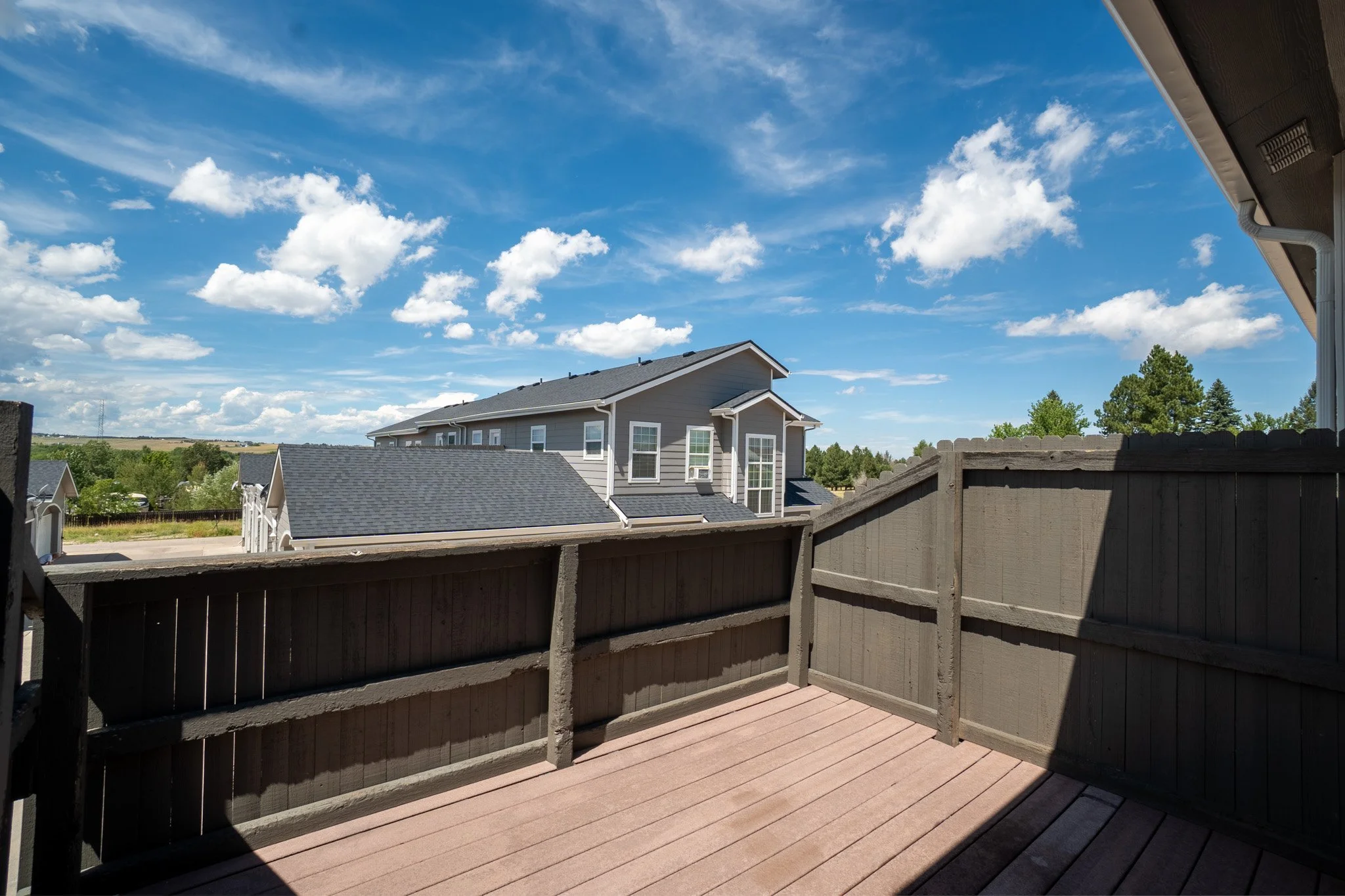 View from a balcony showing neighboring houses, a wide blue sky with white clouds, trees, and fields in the distance.