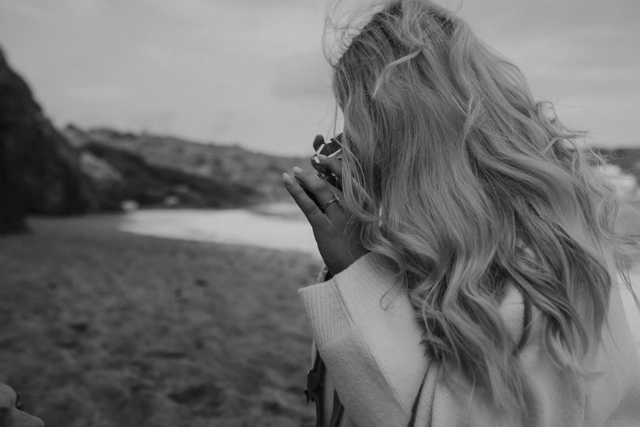 Black-and-white photo of a woman with long hair taking a picture outdoors with a camera, with mountains in the background