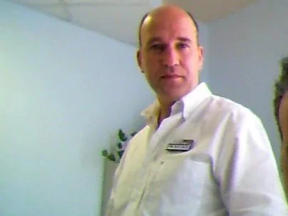 Man in white shirt standing indoors with a light blue wall and plant in the background.