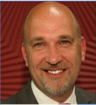 A smiling middle-aged man with a bald head and a gray beard, wearing a suit and tie, standing in front of a red curtain background.