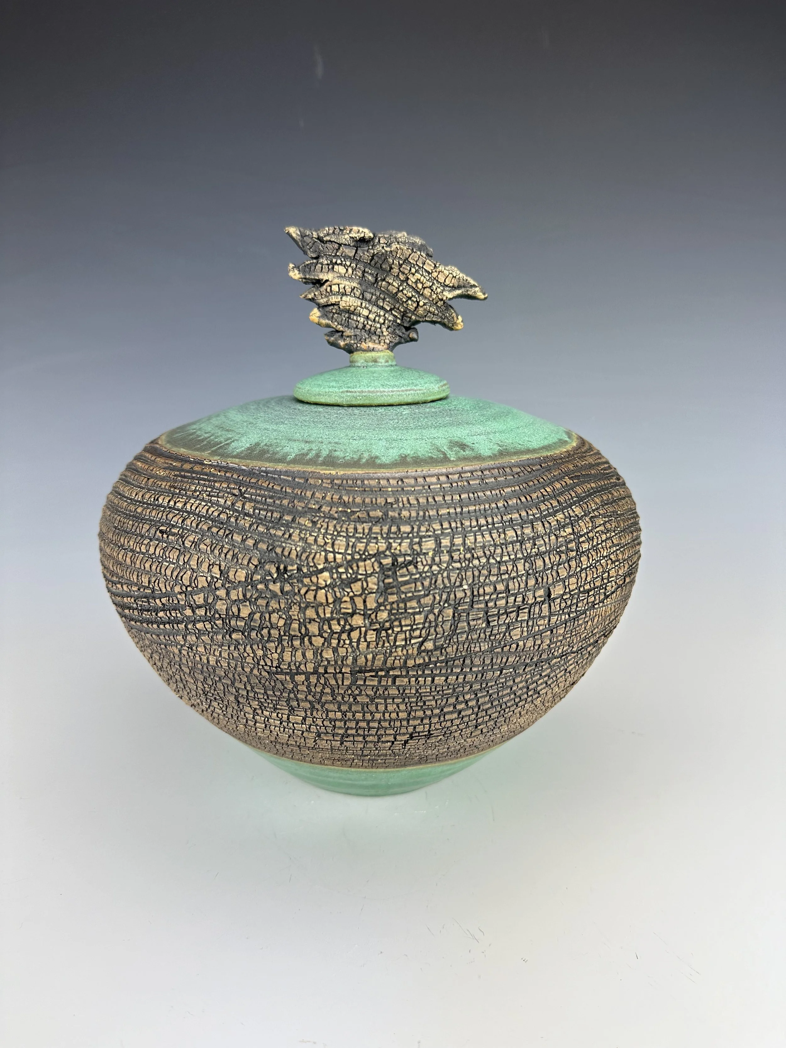 A decorative ceramic jar with a textured, dark, cracked pattern on the body and a greenish glaze on the lid and base. The lid features a tree-shaped handle.