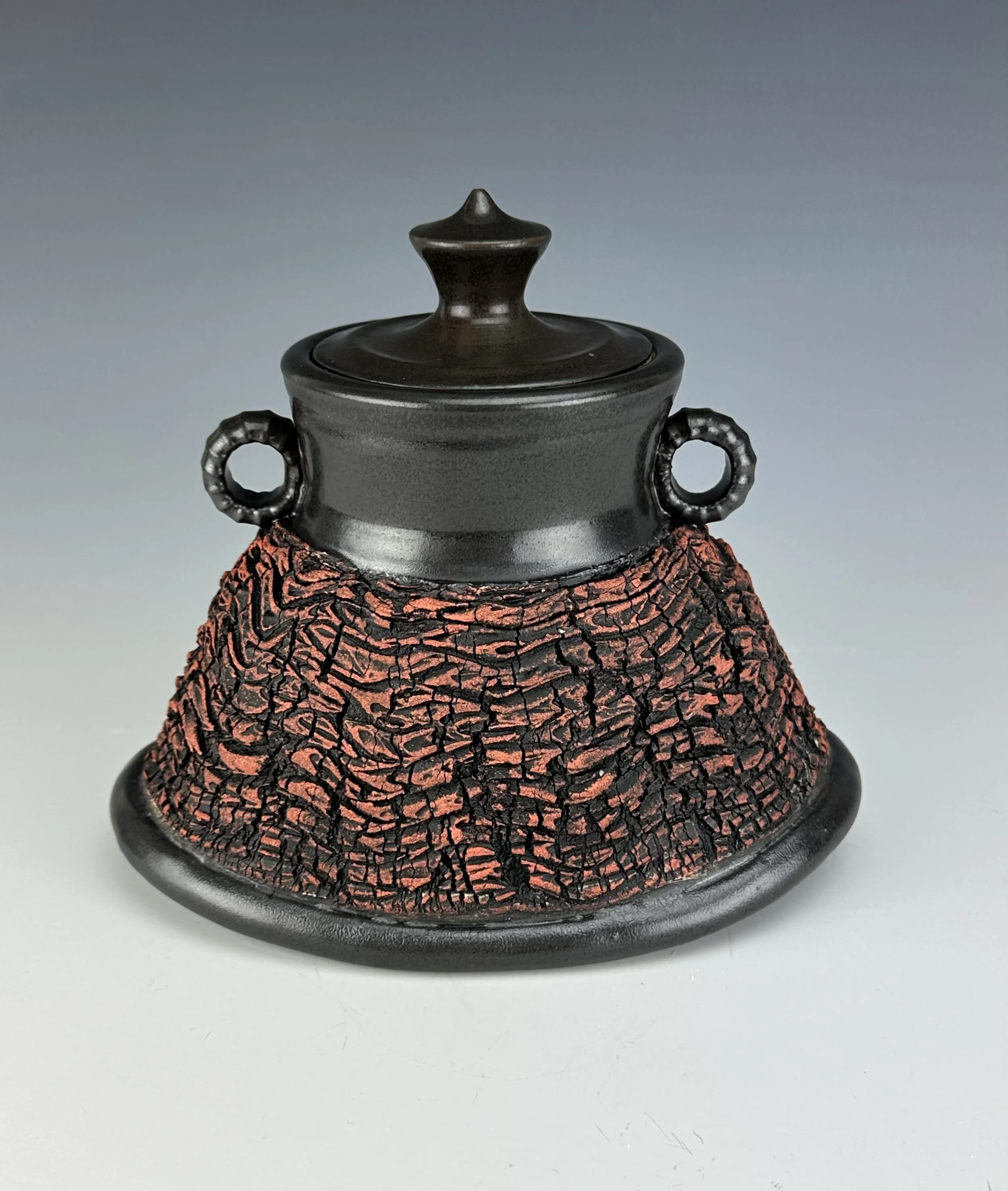 A black and red textured ceramic container with a lid, featuring two circular handles on the sides.