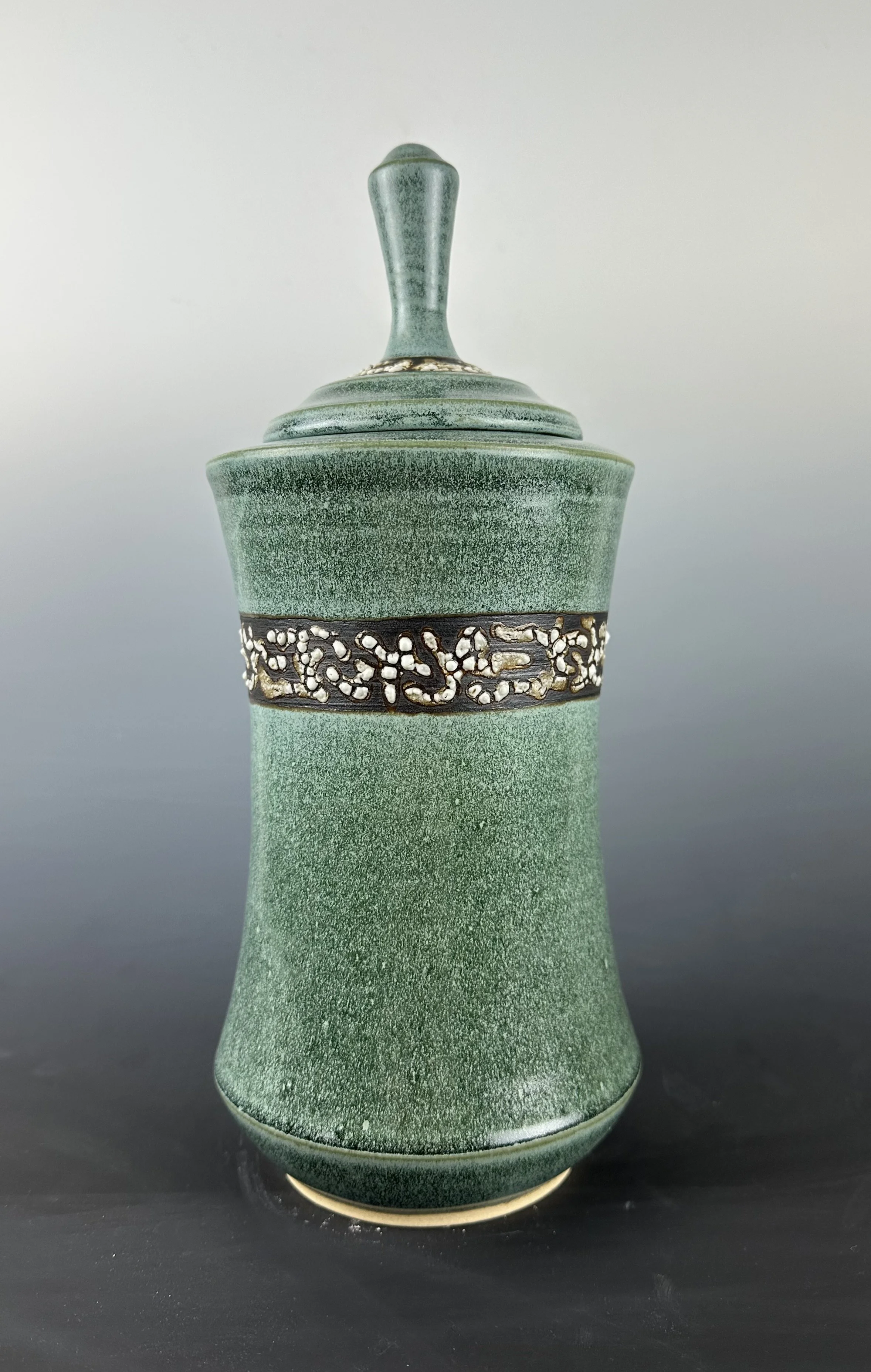 Green ceramic vase with silver and black decorative band near the top, placed on a dark surface against a gray background.