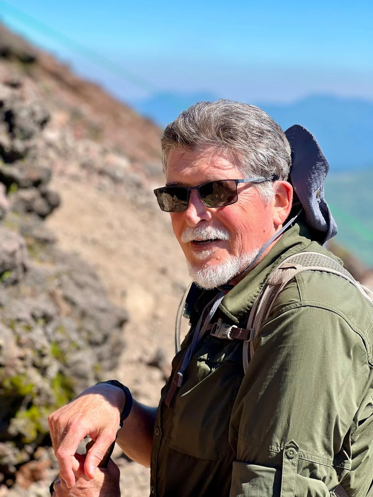 A smiling elderly man with gray hair and a beard, wearing sunglasses, a green jacket, and a backpack, outdoors on a mountain trail with rocks and blue sky in the background.