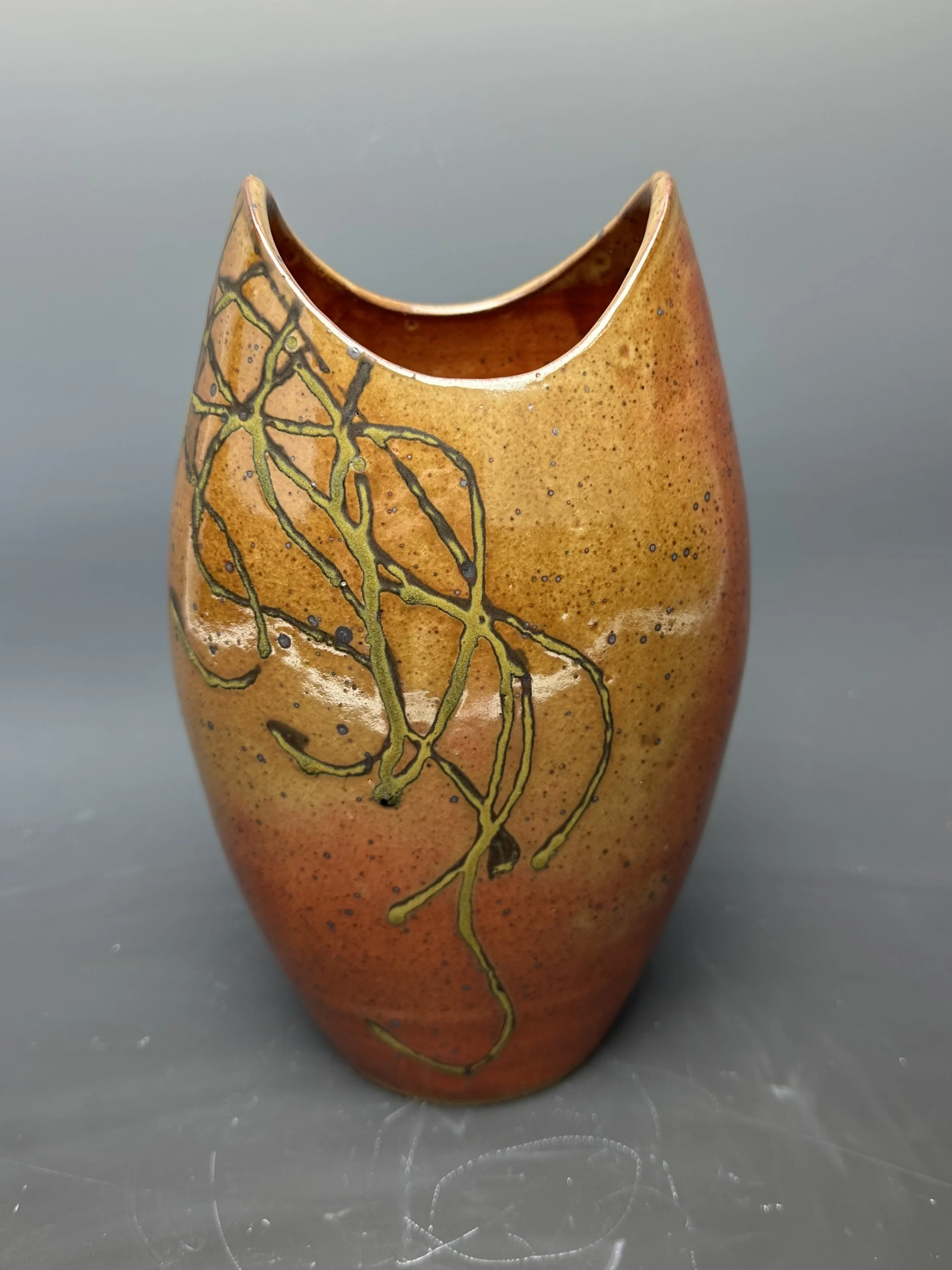 A tall, narrow ceramic vase with a rounded top opening and orange-brown glaze. It features a black and yellow-green abstract linear design resembling a branch or vine across its surface. The background is a plain, muted gray.