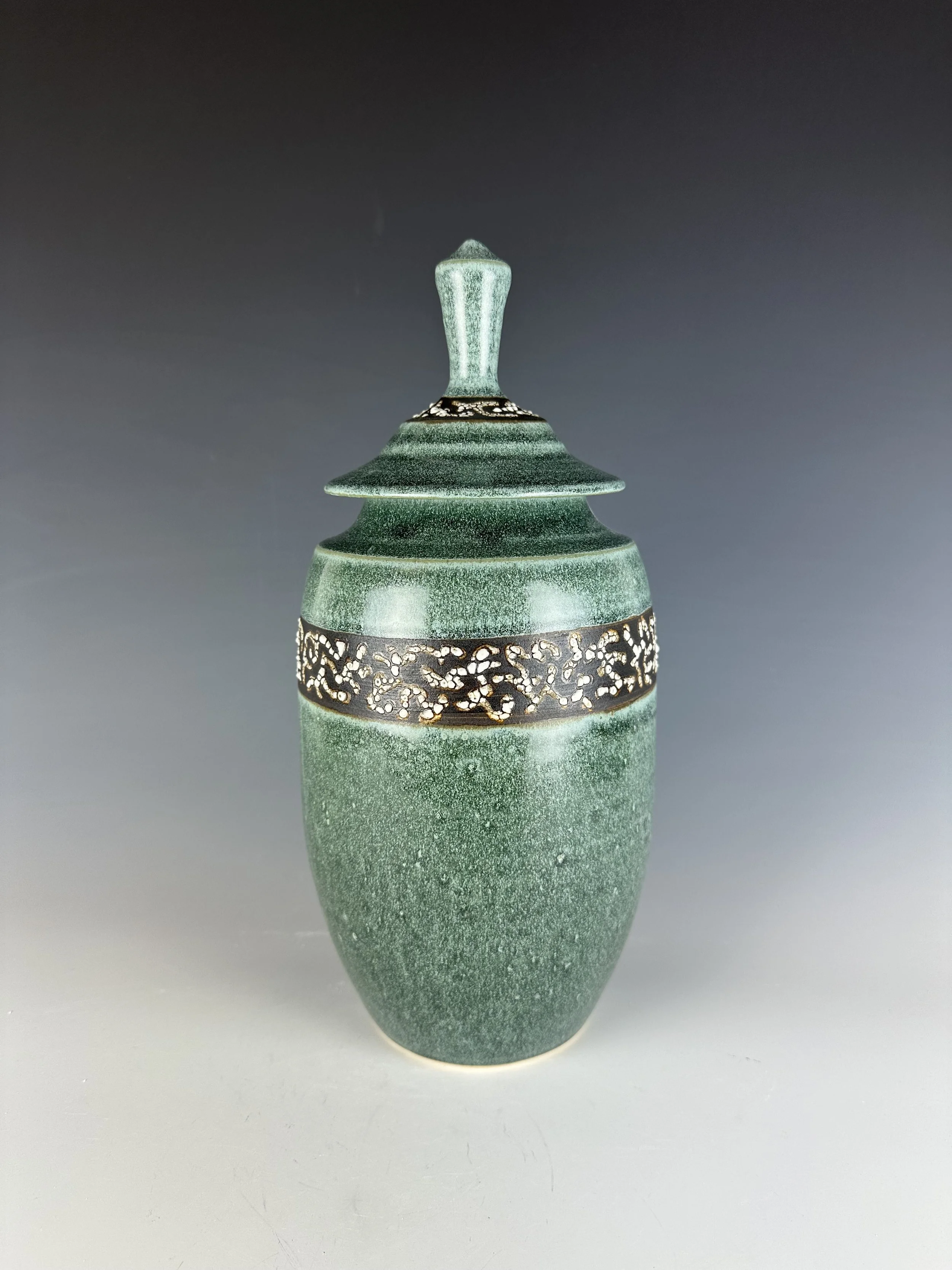 A green ceramic vase with intricate black and white decorative patterns around its middle, featuring a textured surface, placed on a plain surface with a gradient background.