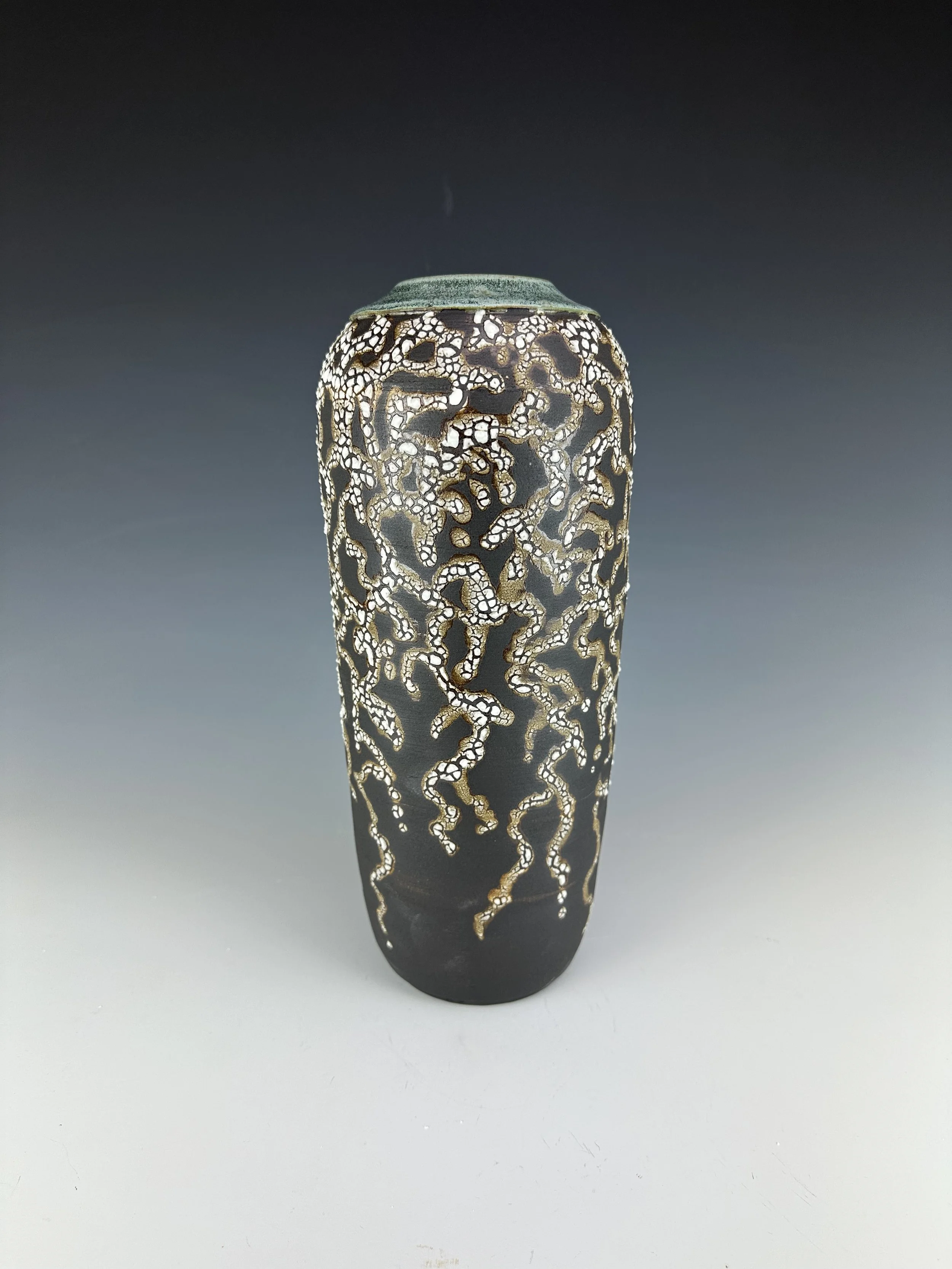 A tall, cylindrical object with an intricate, lace-like pattern in black, white, and gold colors against a gradient gray background.