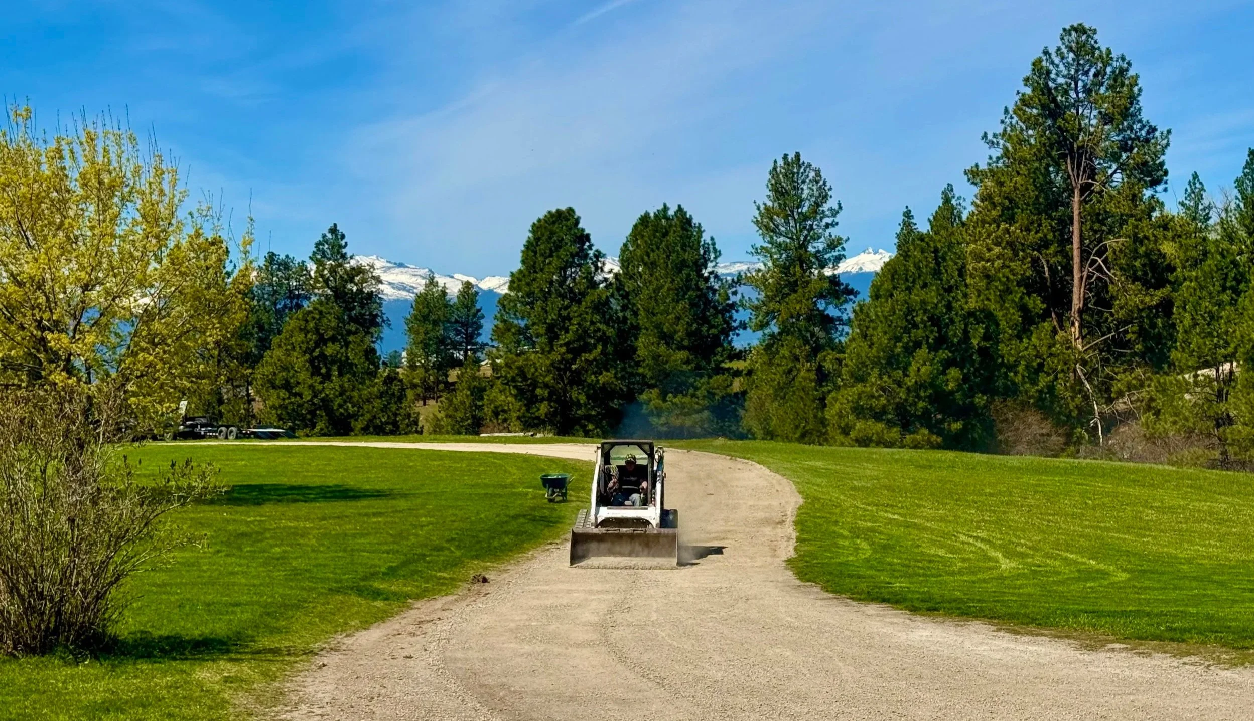 A golf course with a dirt path, a person driving a small vehicle, and mountains with snow in the background