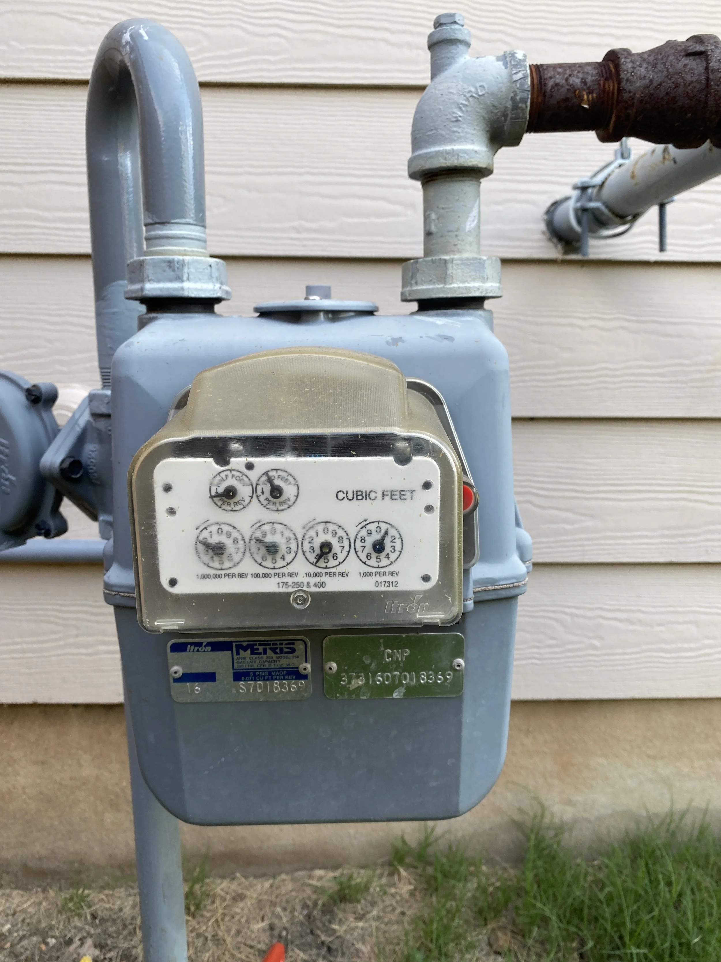 An outdoor gas meter attached to a house with vinyl siding. The meter measures cubic feet of gas usage with dial gauges and is mounted on a metal post. There is some moss or rust on the pipe connection.