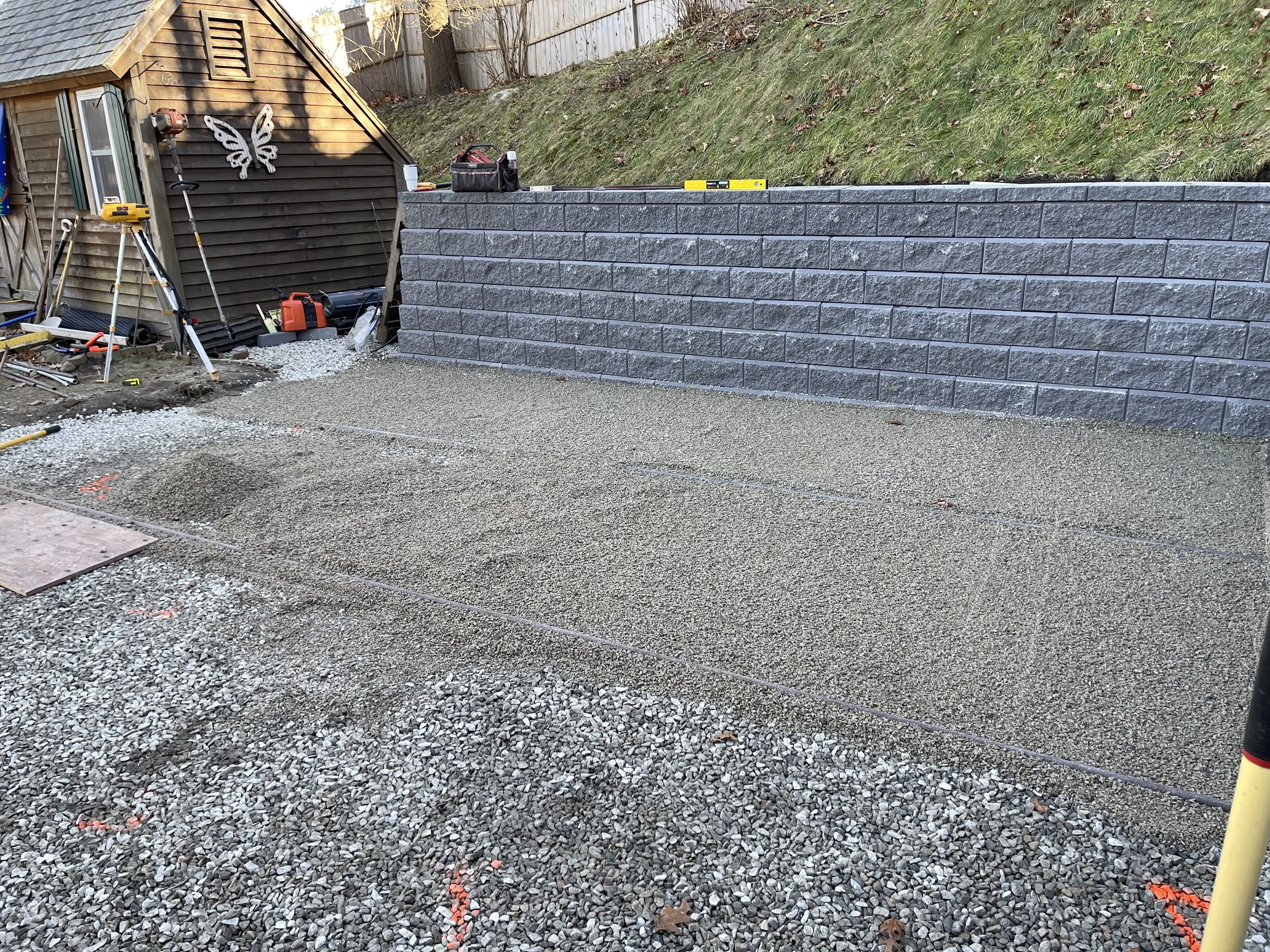 Construction site with gravel ground, a gray stone retaining wall, and a small wooden shed with butterfly decoration. Tools and construction equipment are scattered around.