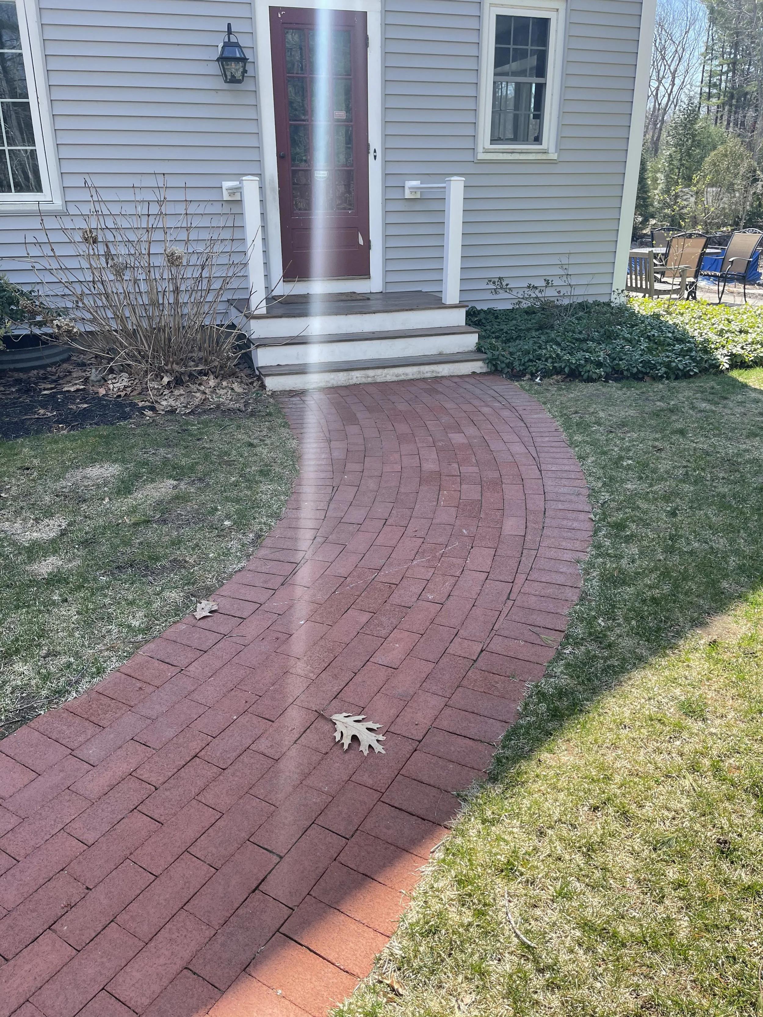 Red brick curved walkway leading to a house entrance with stairs, surrounded by grass and bushes, with a latch on the door, window on each side, and outdoor chairs in the background.