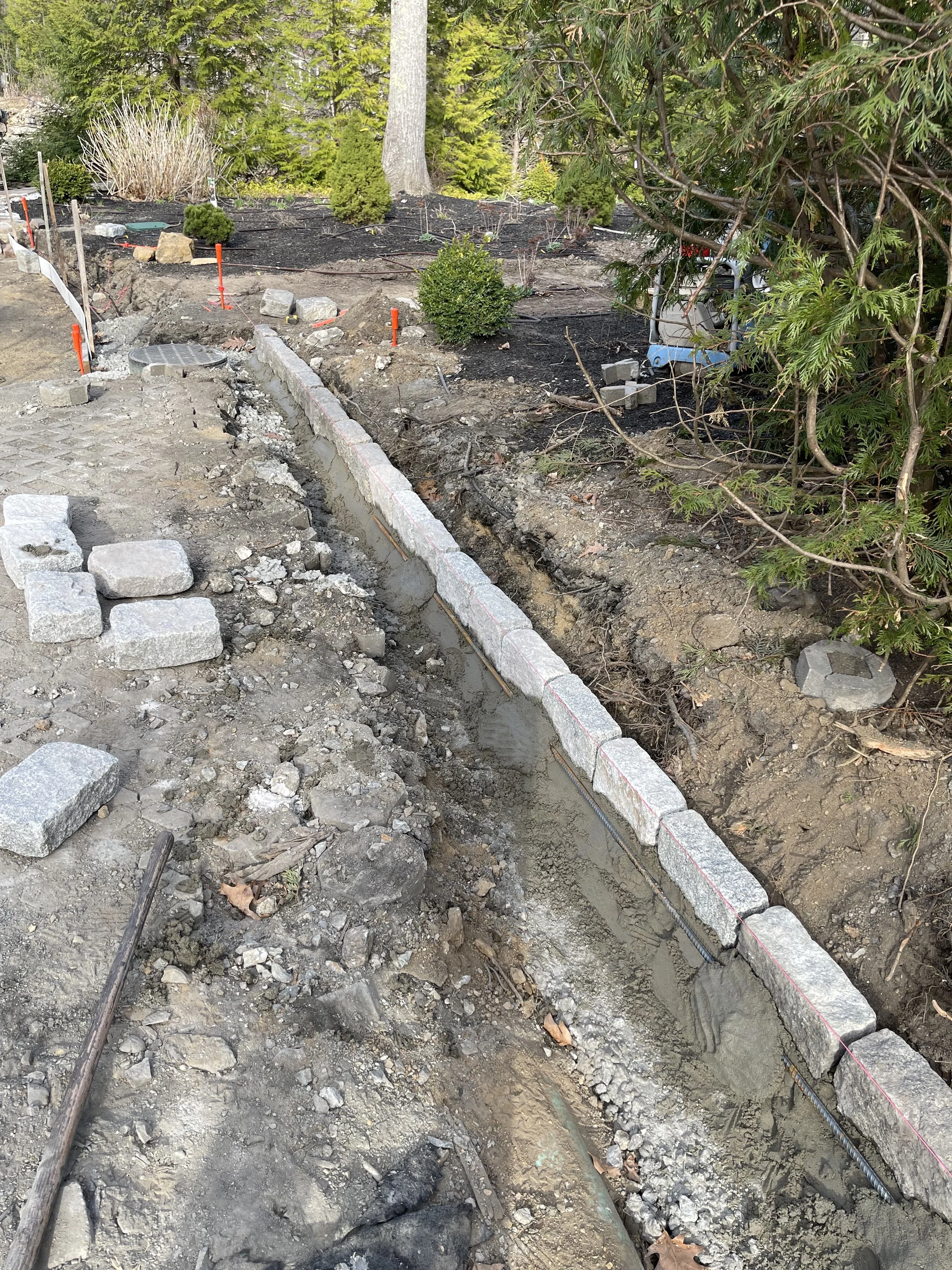 Construction site with a partially built stone wall, dirt and rocks, and some small plants in the background.