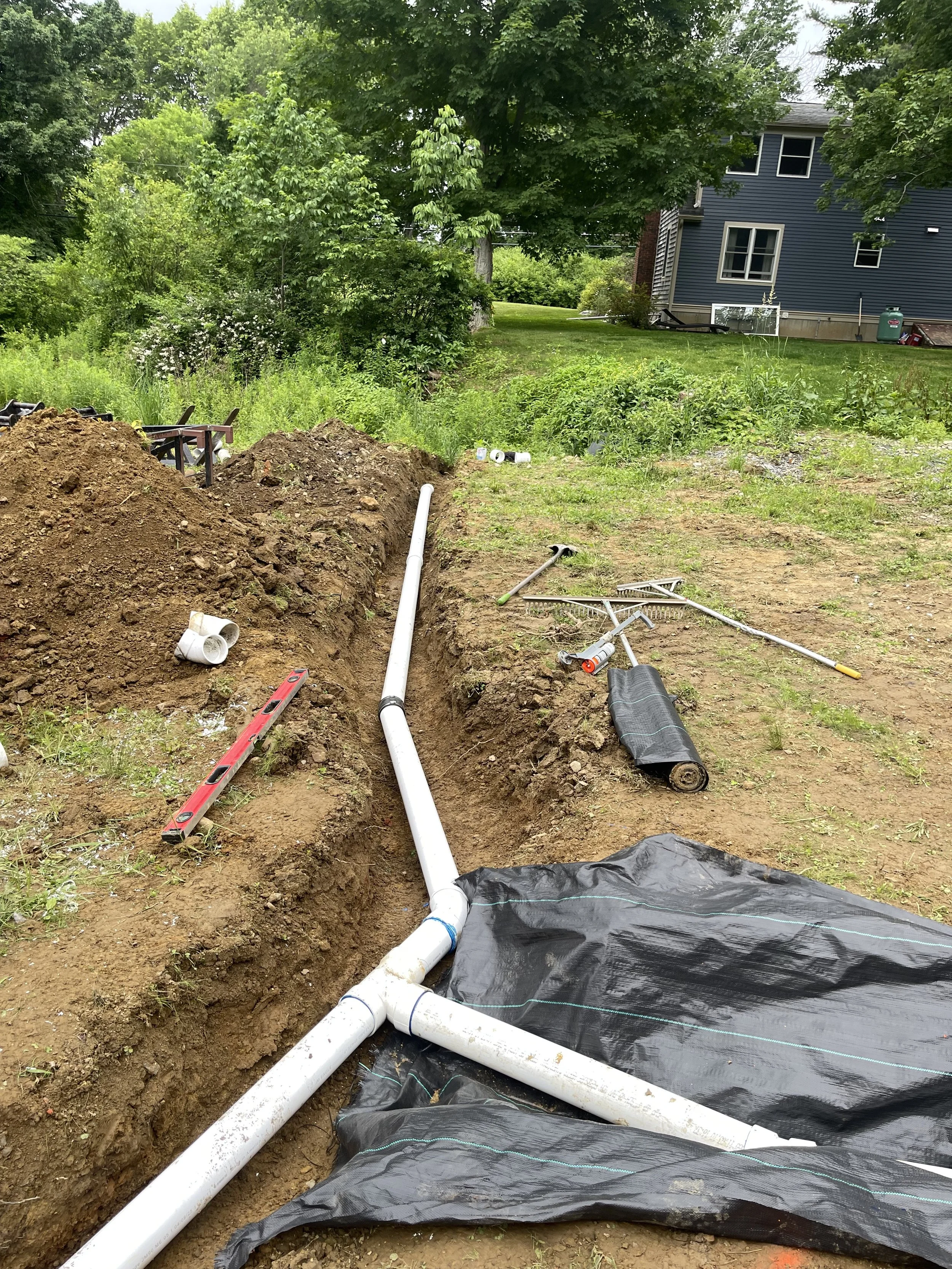 Construction site with a trench dug in the ground for a French drain installing underground piping, with white PVC pipes, a level, a rake, and a black drainage line layer visible, surrounded by grass, trees, and a house in the background.