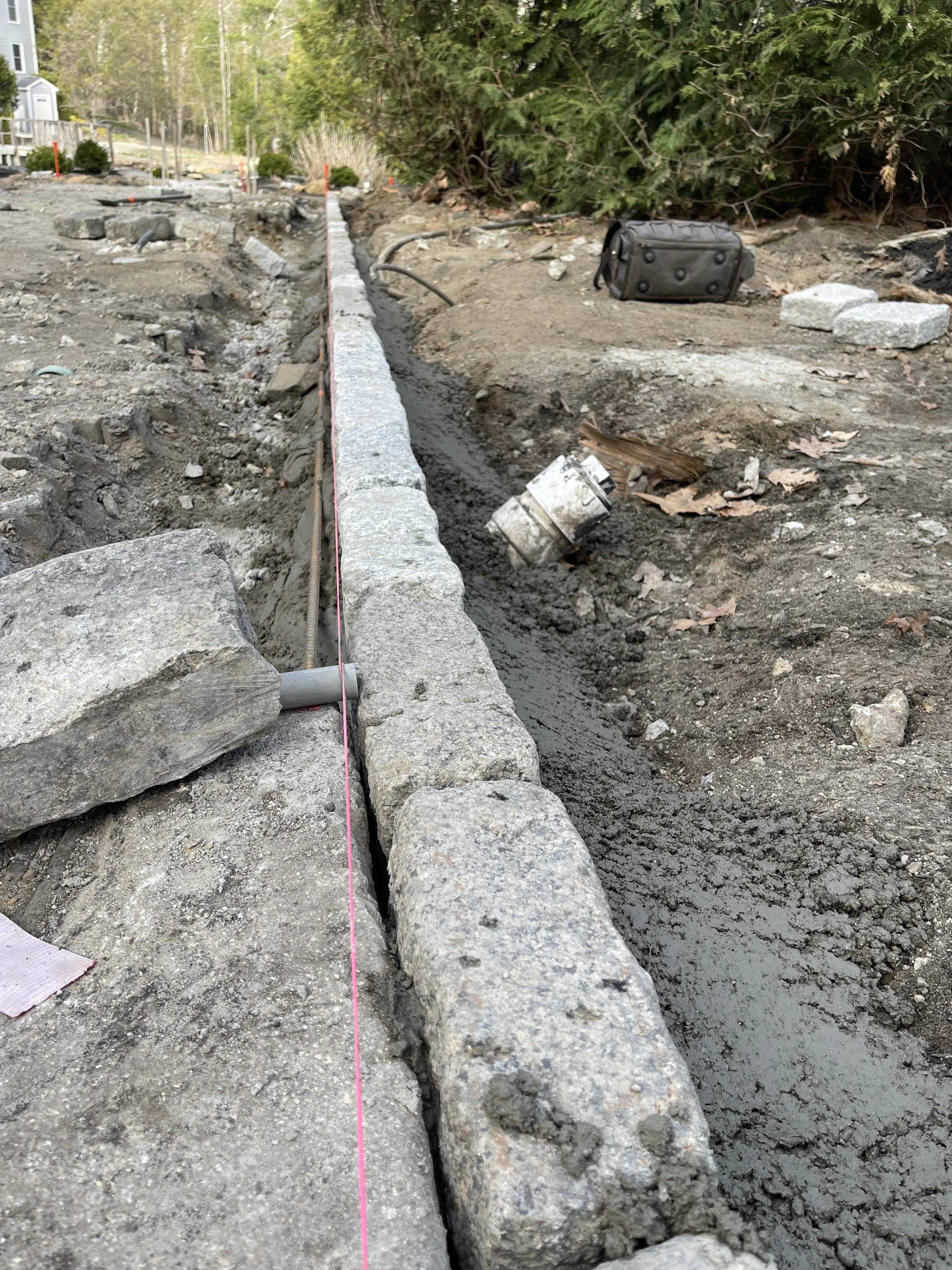 Construction site with a stone border, black bag, and construction tools, with dirt and trees in the background.