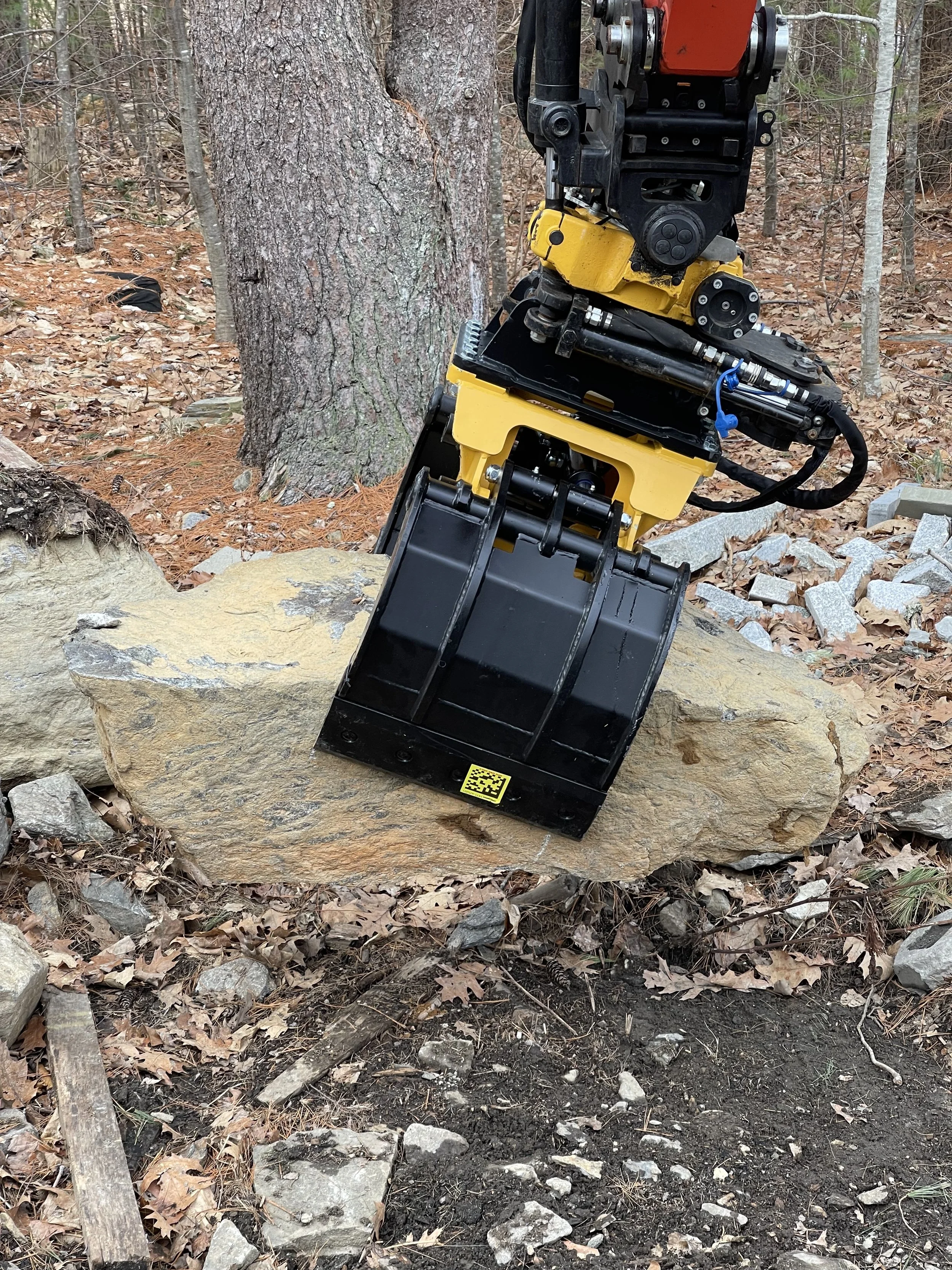 A piece of heavy machinery or robotic equipment with orange, black, and yellow parts is positioned next to a large tree in a forest setting. The machine is on rocky and leaf-covered ground.