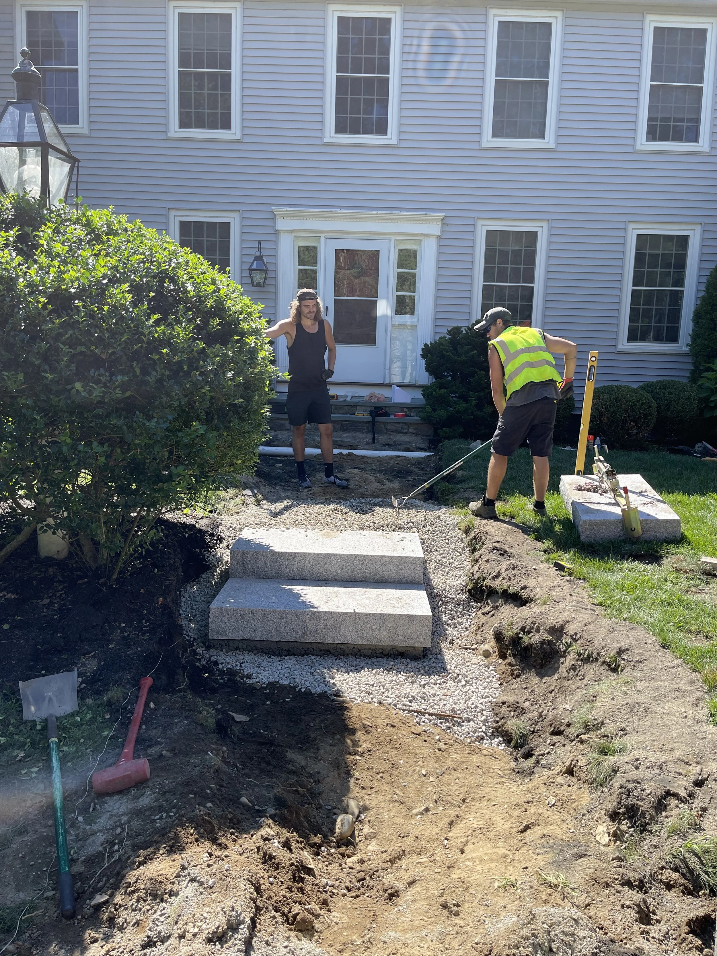 Two men working on a landscape project in front of a house with white siding and multiple windows. They are working on a pathway with concrete steps and gravel. Tools and materials are scattered around, including shovels and a level.