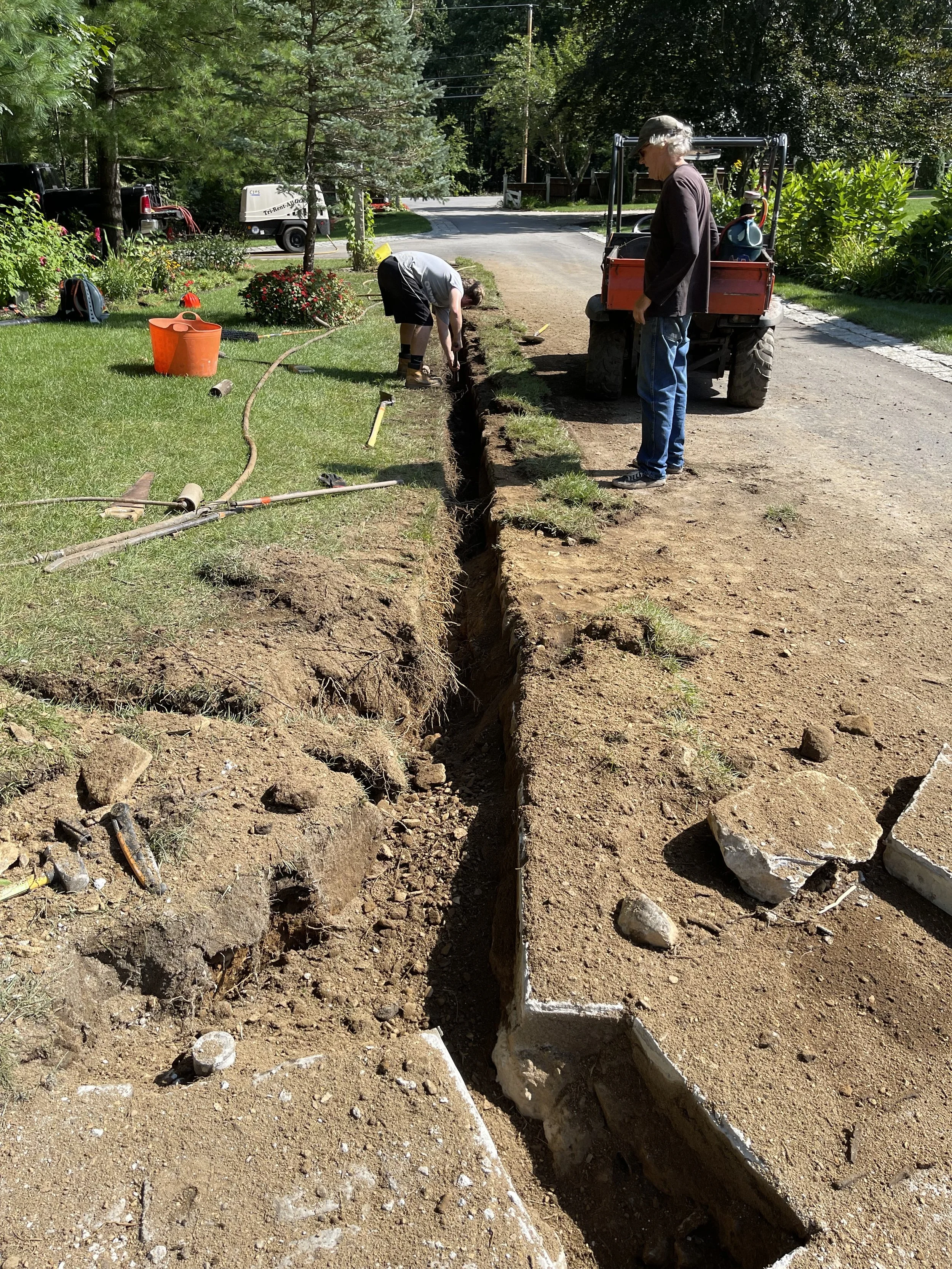 Two men are working on a trench in a front yard, surrounded by tools, rocks, and a small orange utility vehicle. One man is bending down, operating the trench, while the other stands nearby. The backyard has trees, plants, and a driveway, with equipm