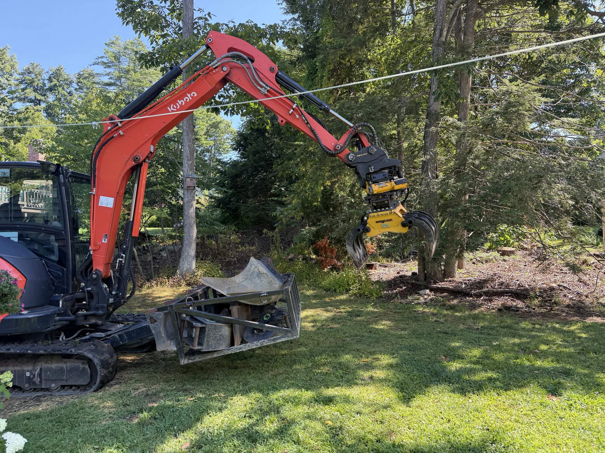 A red compact excavator with a hydraulic arm and a grapple attachment, working on a hillside surrounded by trees, with a blue sky overhead.