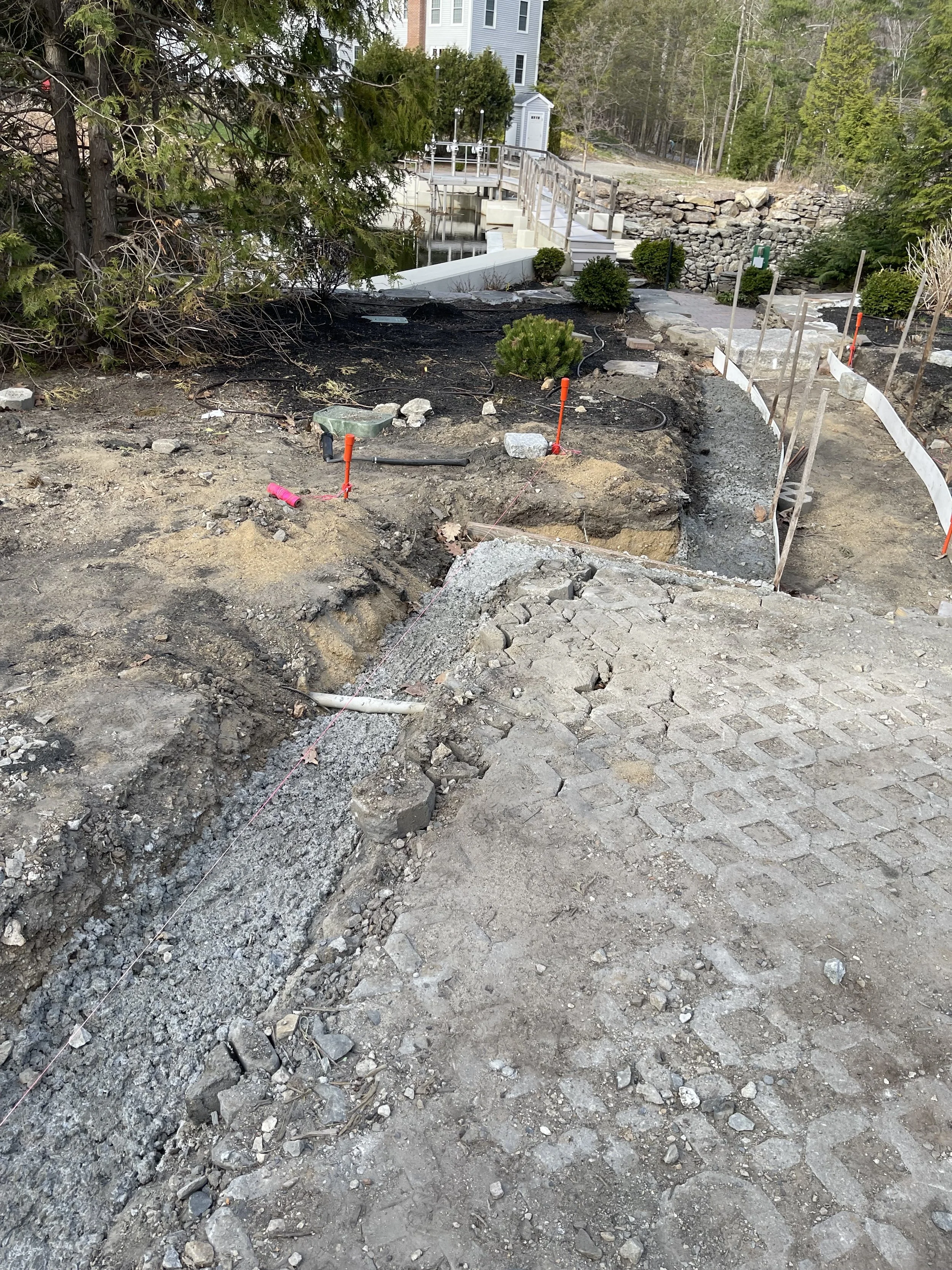 A construction site showing a dirt and gravel pathway with excavation work, orange markers, and construction tools. In the background are trees, shrubs, a stone wall, and a white house with a deck and stairs.