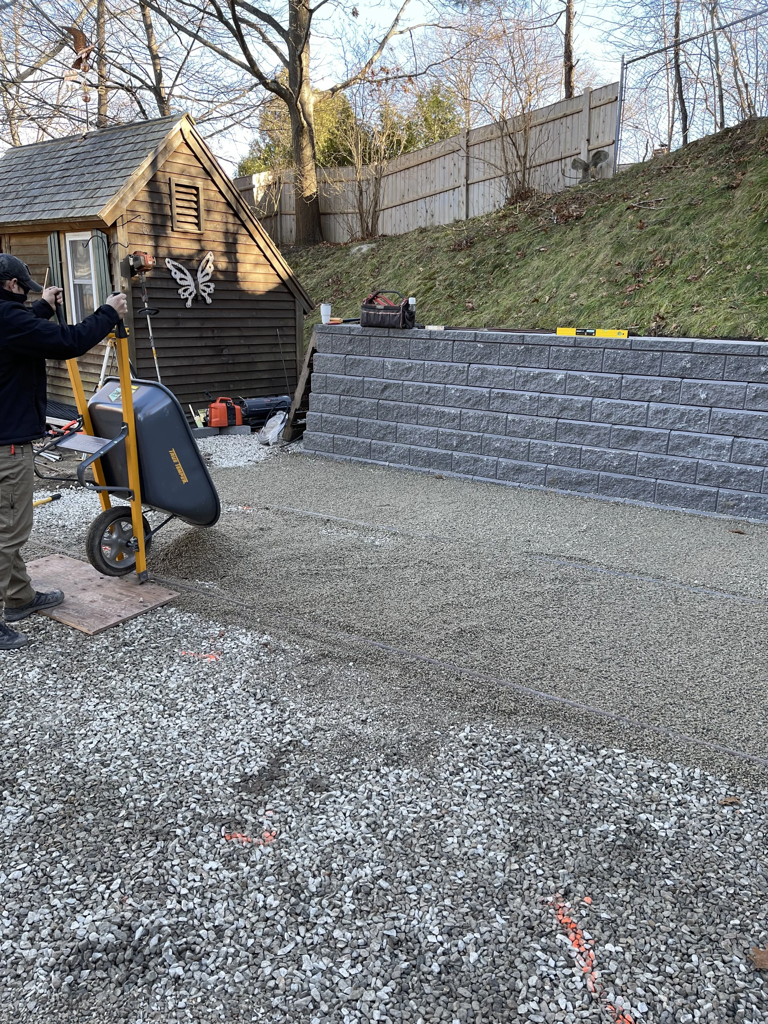Workers are laying down gravel on a construction site, with a gray stone retaining wall and a wooden shed in the background.
