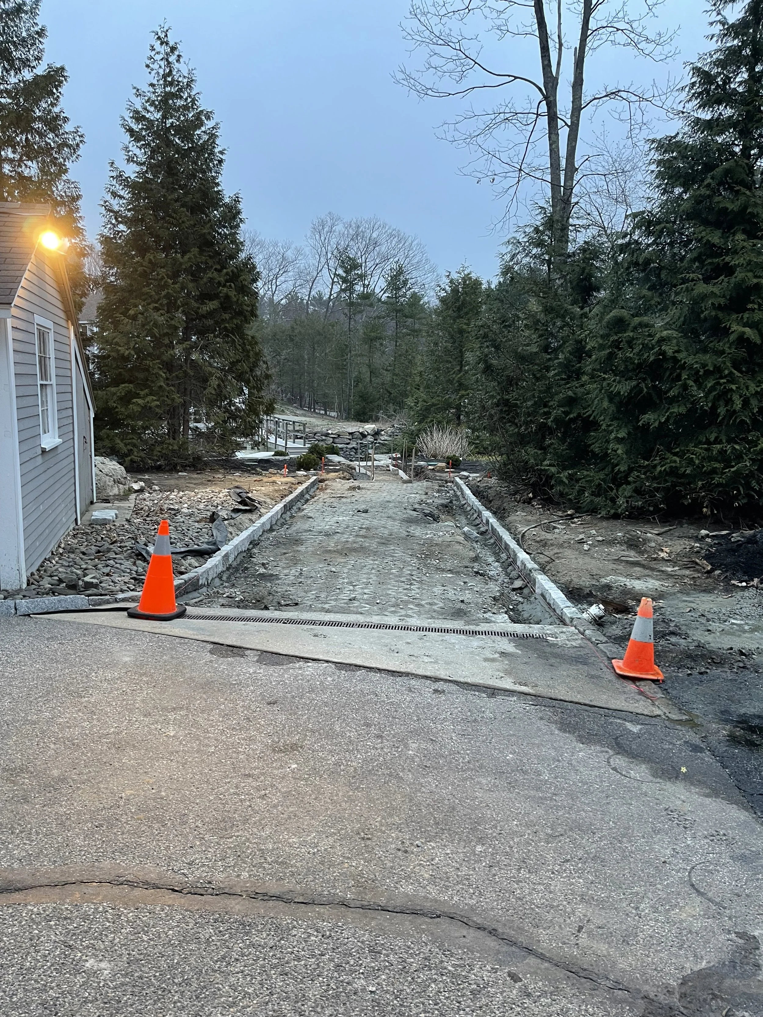 A road under construction with orange cones marking the area, surrounded by trees and a house on the left side, with a rocky pathway ahead and a wooded hill in the background.