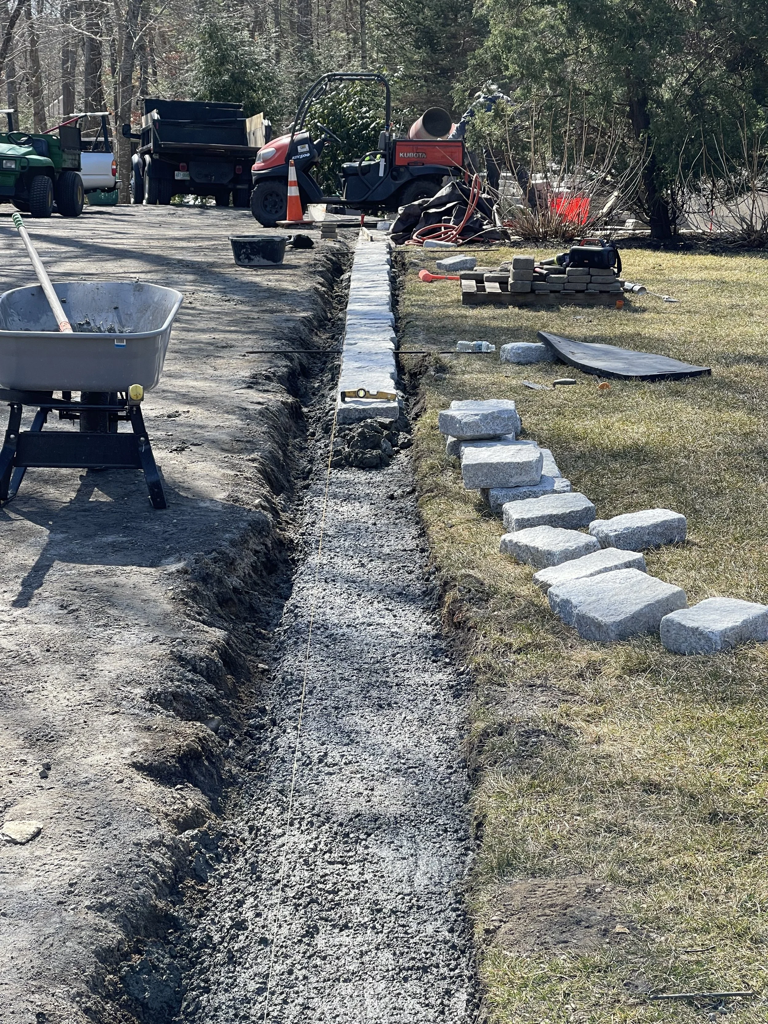 Construction site with a trench being dug along a gravel pathway, surrounded by construction tools, stacked bricks, and parked utility vehicles in a wooded area.