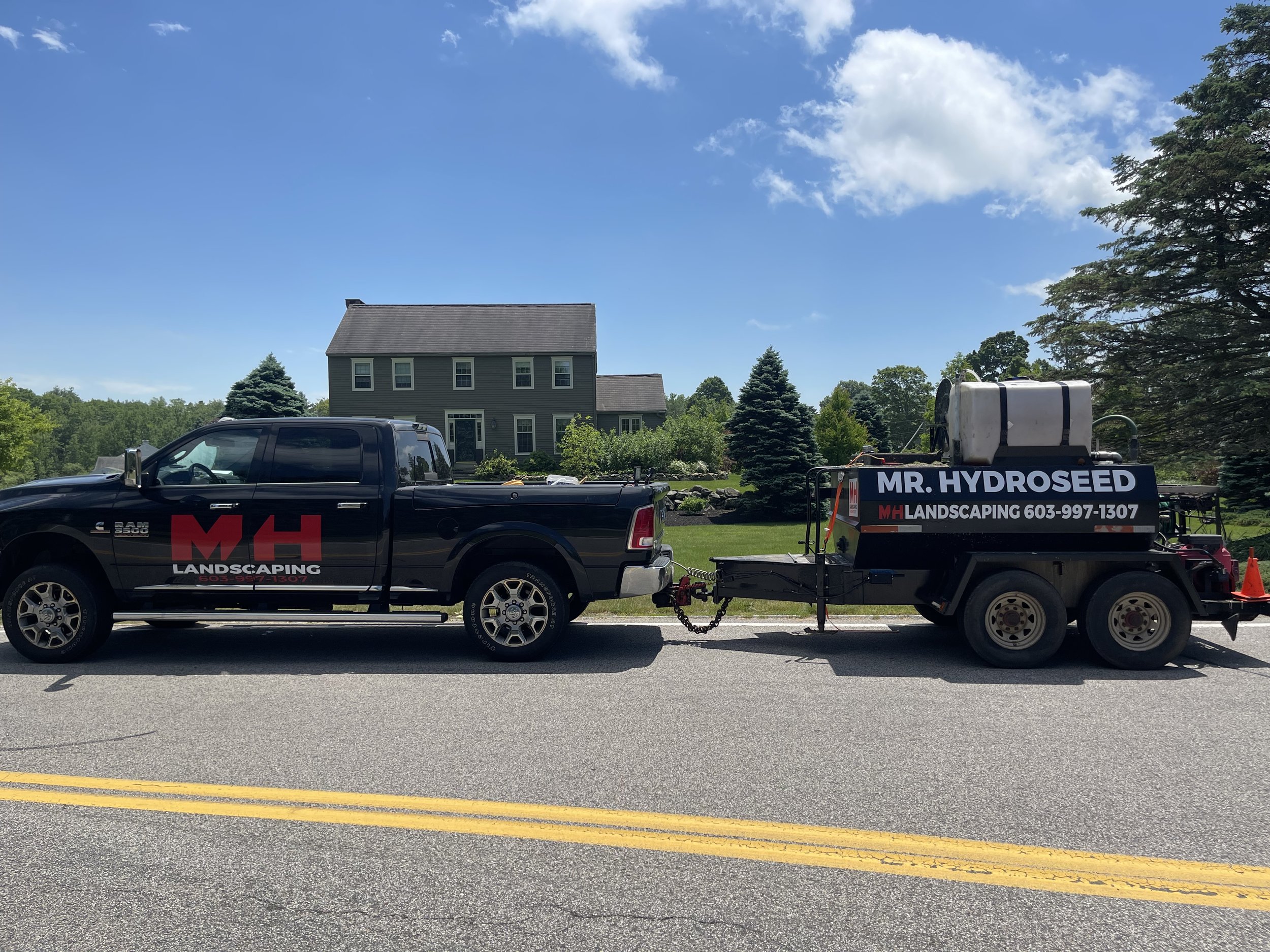 A black pickup truck with red and white branding for MH Landscaping is parked on a road, towing a landscaping spray machine labeled 'Mr. Hydroseed.' In the background, there's a green house with trees and a bright blue sky with a few clouds.