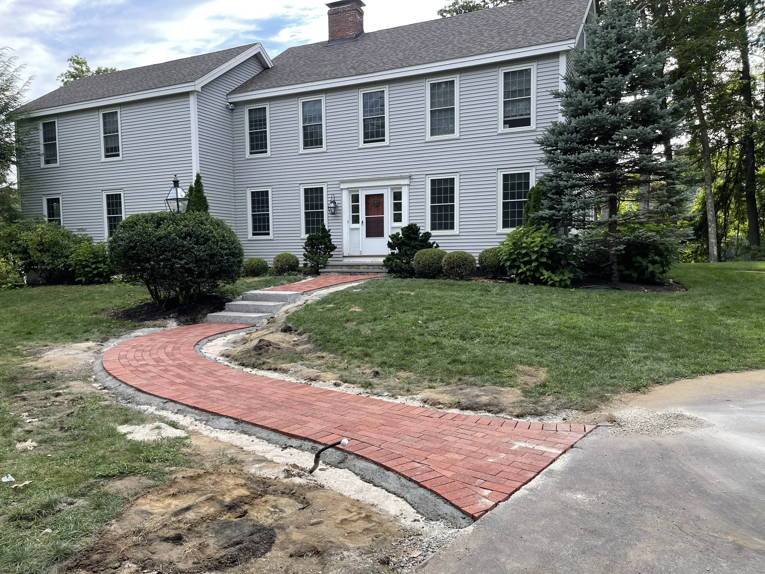 A house with white siding and multiple windows, with a brick chimney, surrounded by trees and shrubbery. A new red brick pathway is being constructed in the front yard.