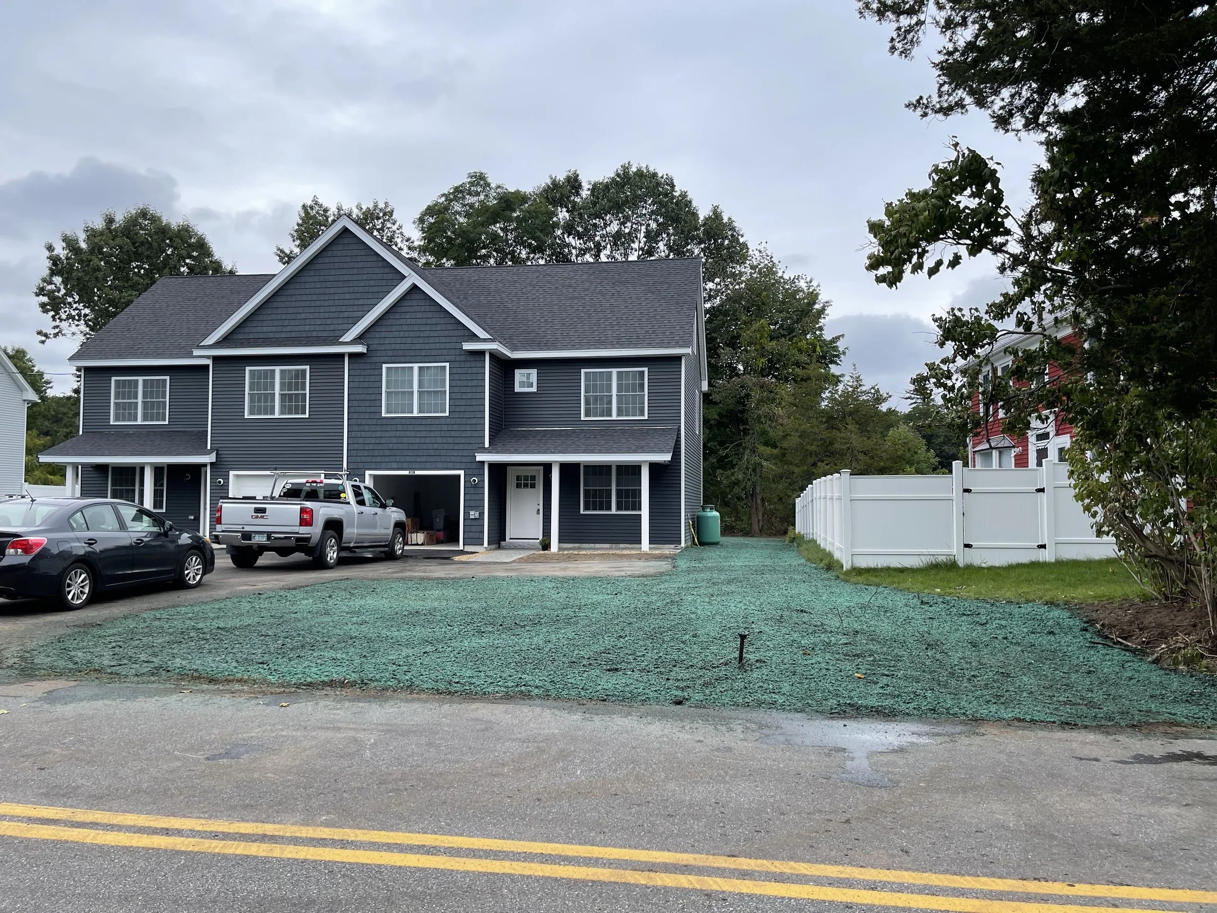 View of a house with blue siding and white trim, multiple cars parked in the driveway, newly hydroseeded yard, trees in the background, and a cloudy sky overhead.