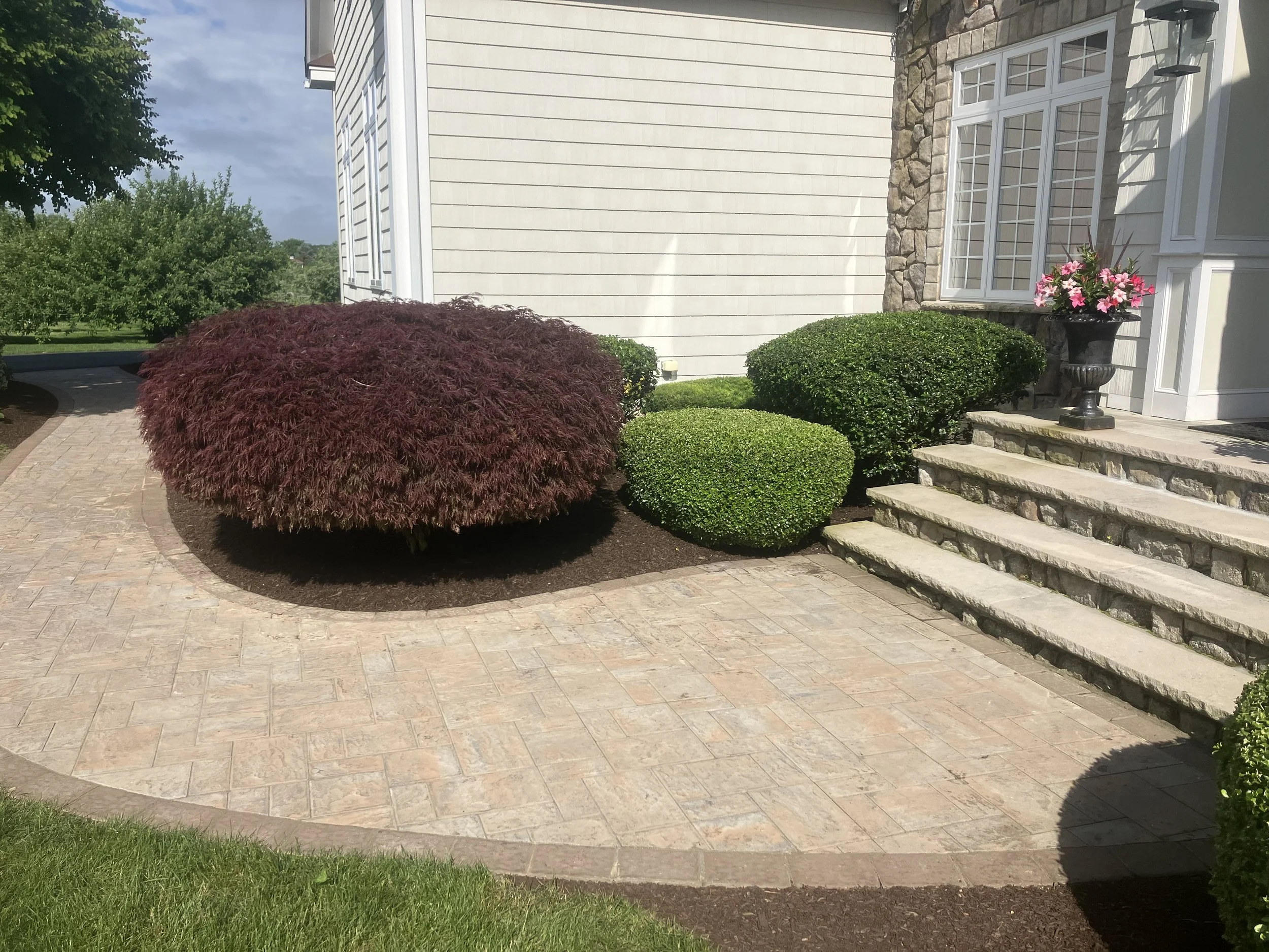 A well-maintained front yard with a curved brick pathway, a large red shrub, neatly trimmed green bushes, a stone planter with pink flowers, and steps leading to the house with white siding and a stone accent wall.