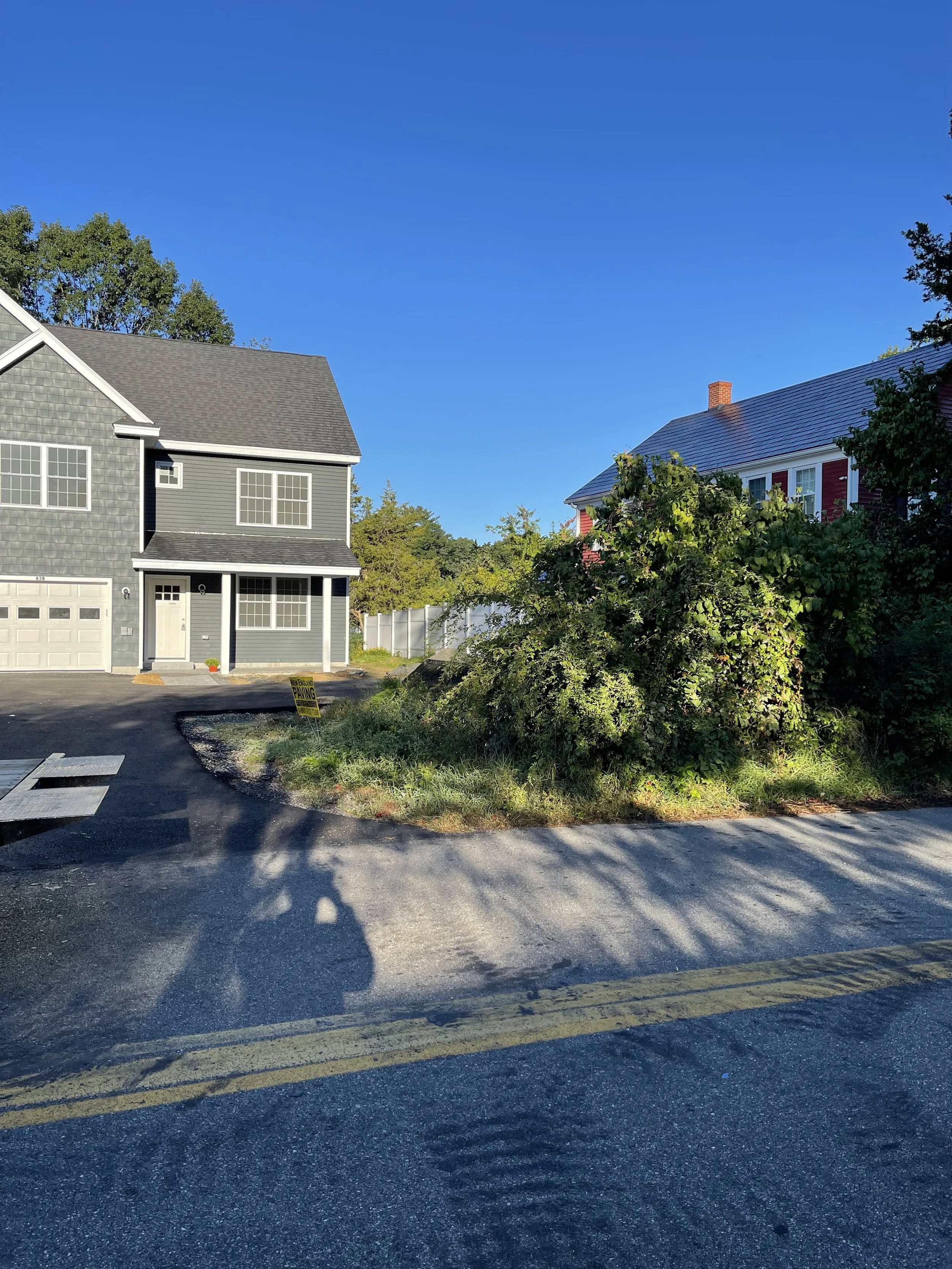 A residential neighborhood with two houses, one gray and one red, under a clear blue sky. The gray house has a garage and front porch, while the red house is partially obscured by trees and shrubs. There is a yellow sign on the ground that says 'No P