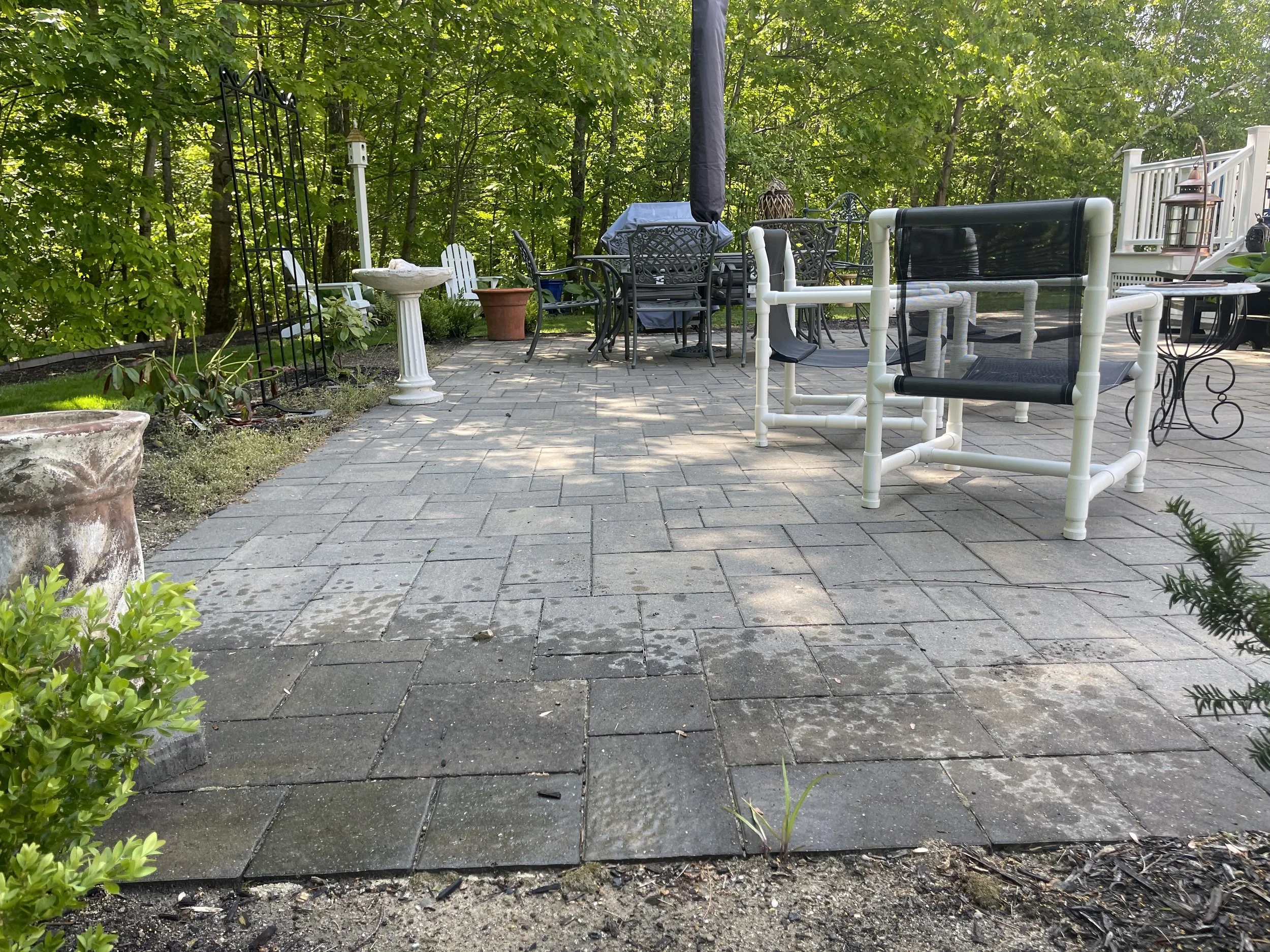 A backyard patio with paver stones, surrounded by lush green trees and bushes. The patio has outdoor furniture including a table with chairs, some white and some black, and decorative items like lanterns. There are plants and garden ornaments along t