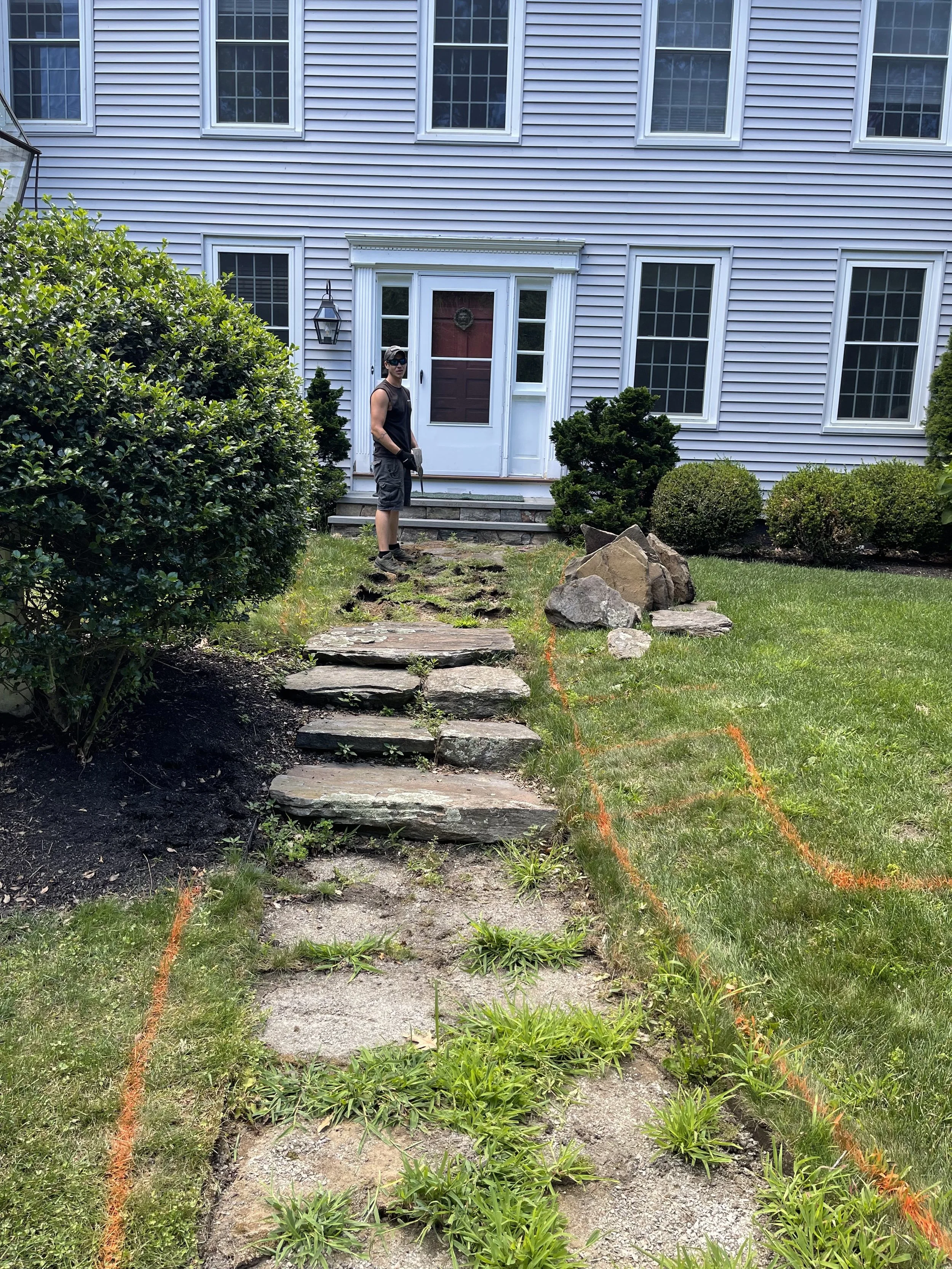 A person standing near a house with a white siding exterior, black window frames, and a red front door. They are wearing sunglasses, a black sleeveless shirt, shorts, and gloves. There is a stone pathway under construction with orange spray paint lin