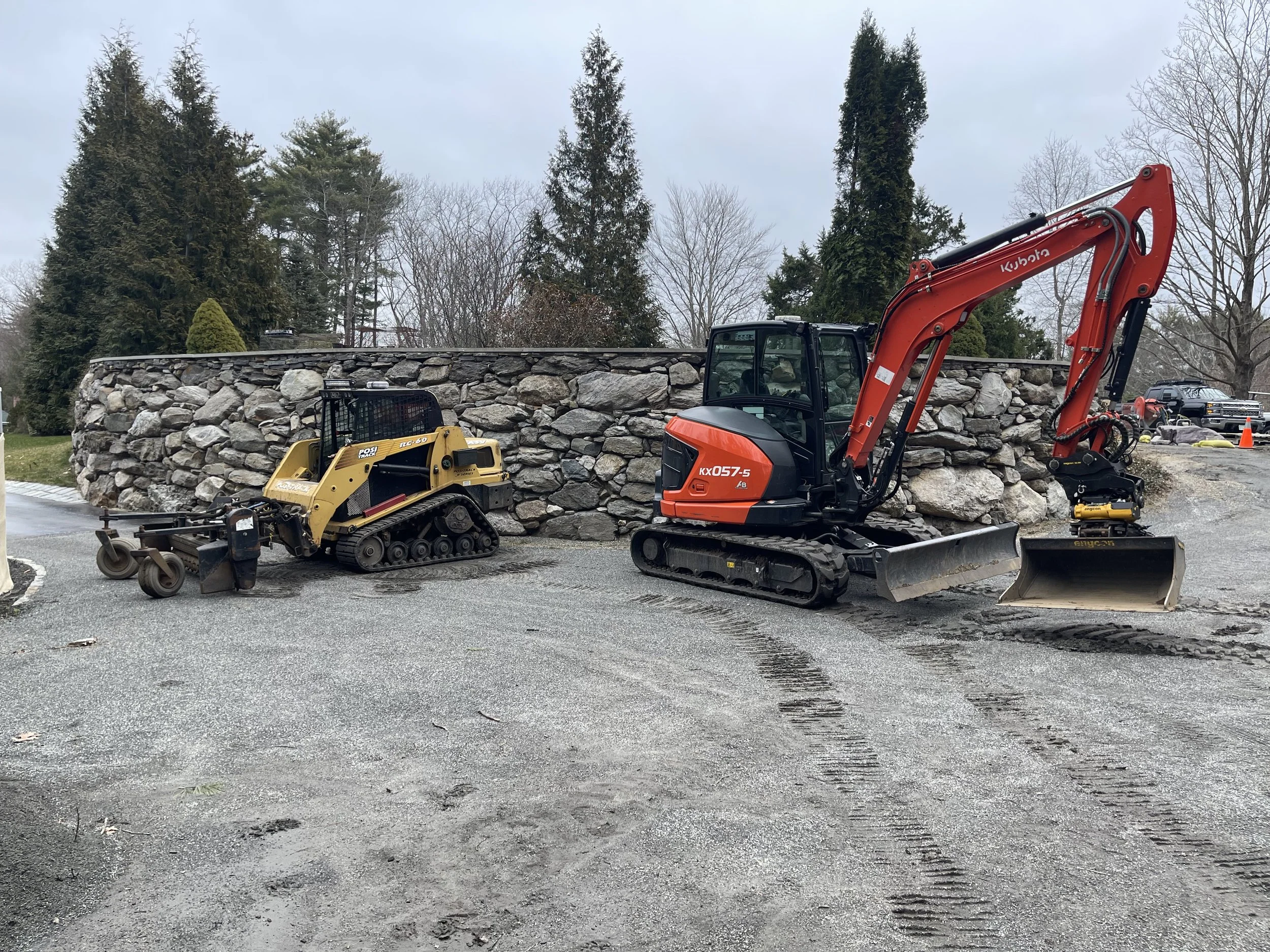 Construction site showing a stone wall with construction equipment and vehicles, including a yellow skid steer with Harley Rake and a larger orange excavator, with trees and homes in the background.