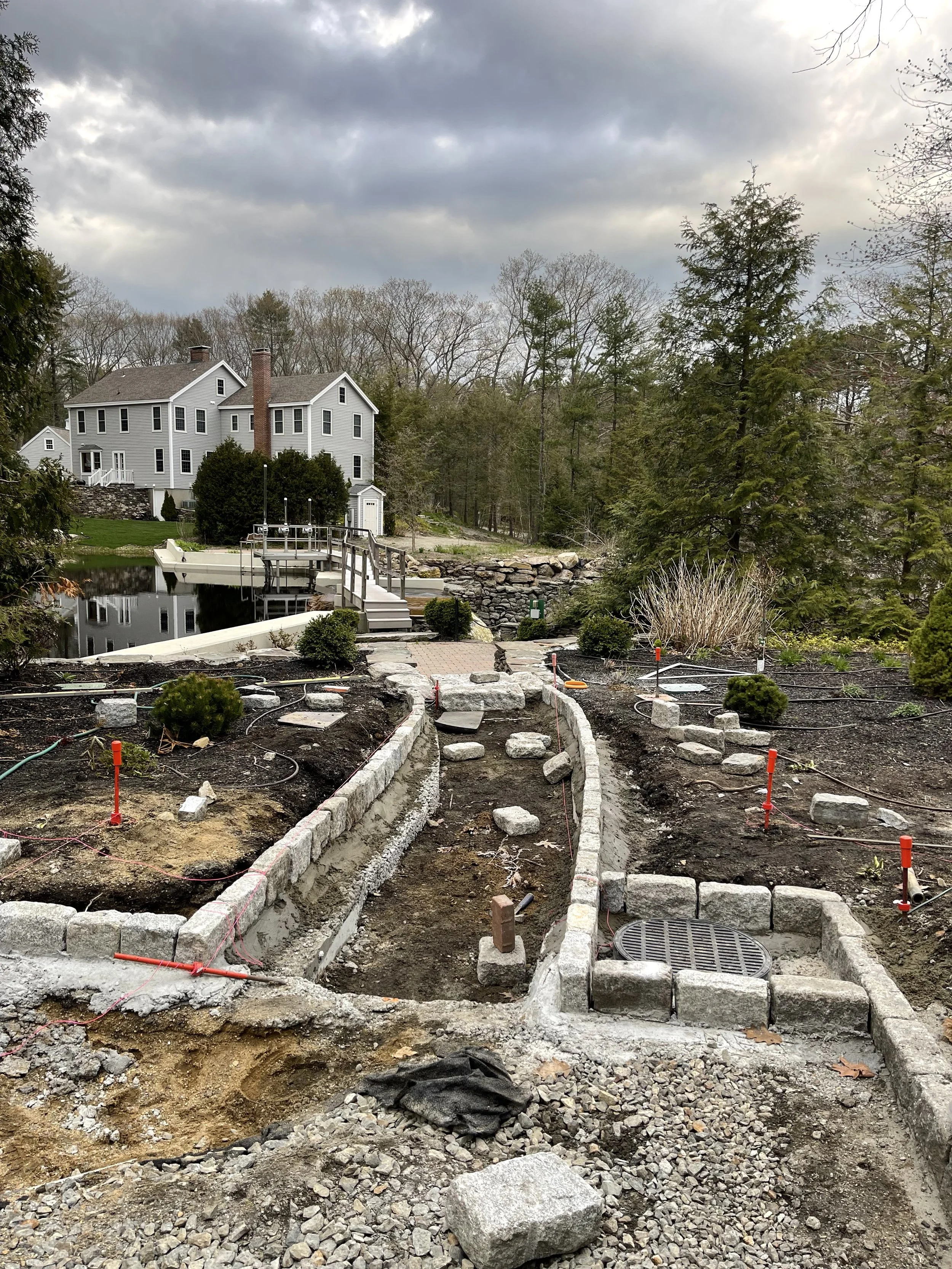 A backyard garden under construction with stone borders and pathways, a house with a pond, and trees in the background under cloudy skies.
